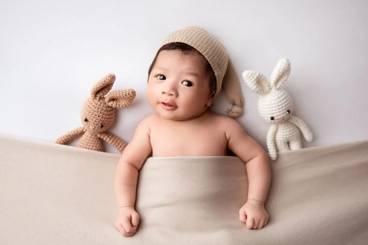 Awake baby in a cream cap between two toy rabbits