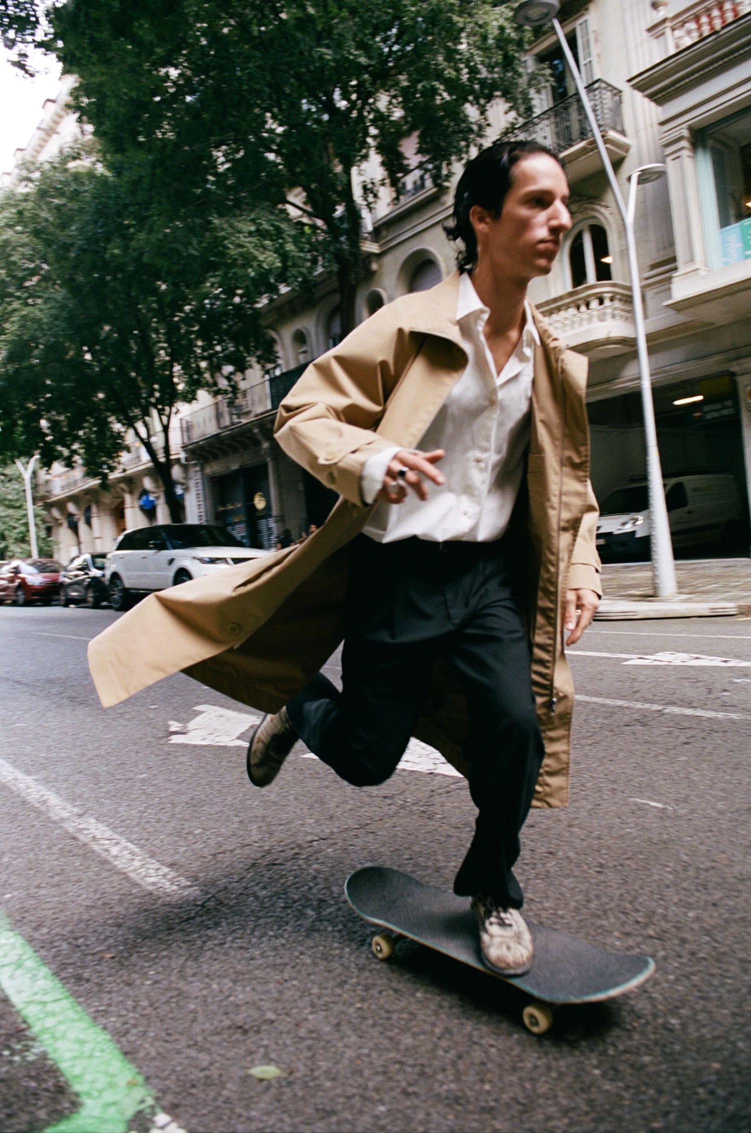 Un skateur à Barcelone, pantalon noir, chemise blanche et trench beige