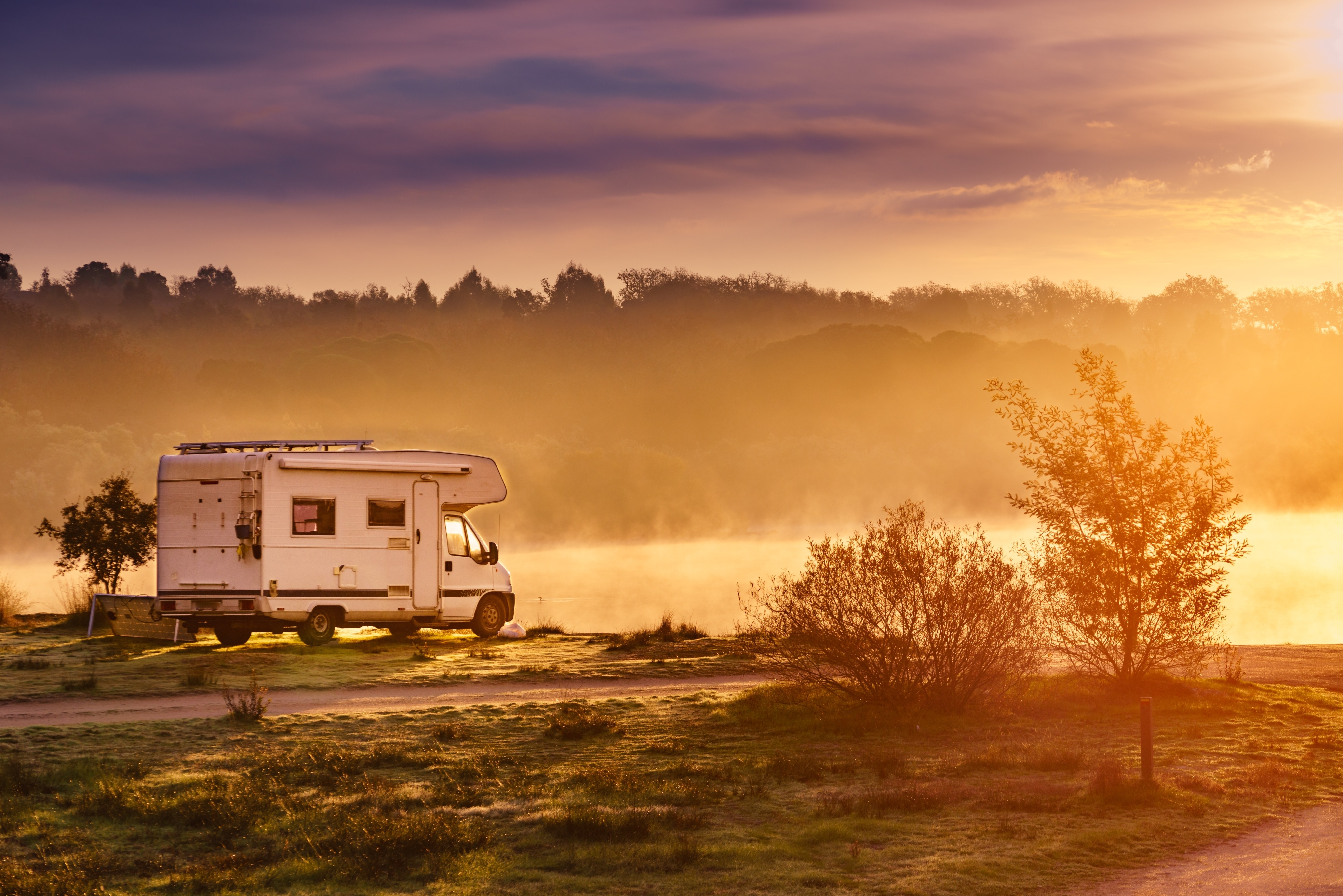 A campervan parked by a misty lake at sunrise, surrounded by trees glowing in warm golden light.