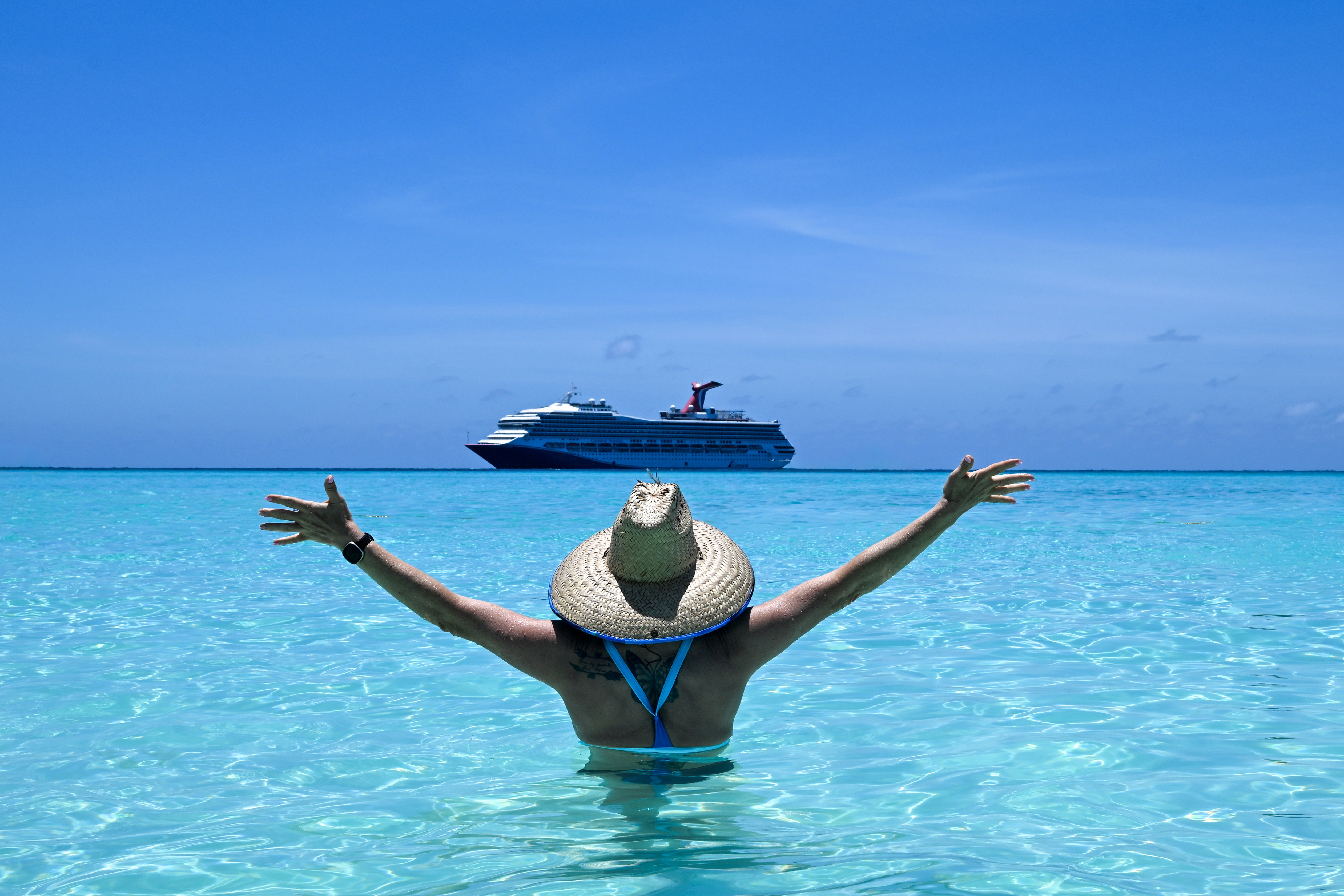 Woman wearing a hat standing in clear blue ocean water looking out at a cruise ship with her arms raised wide