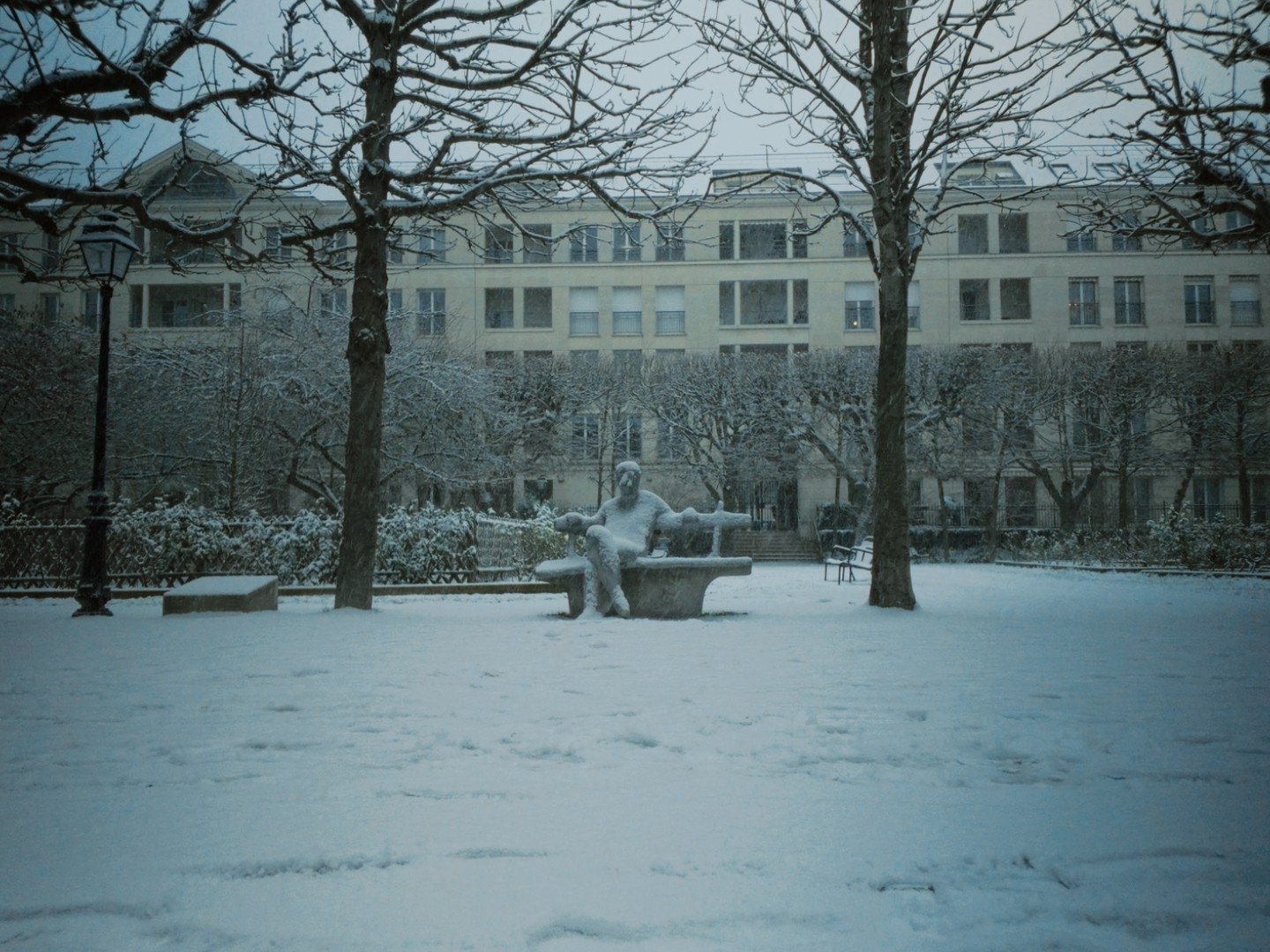Parc enneigé à issy, statue sur banc enneigée, arbres sans feuilles, bâtiments historiques, ciel gris.