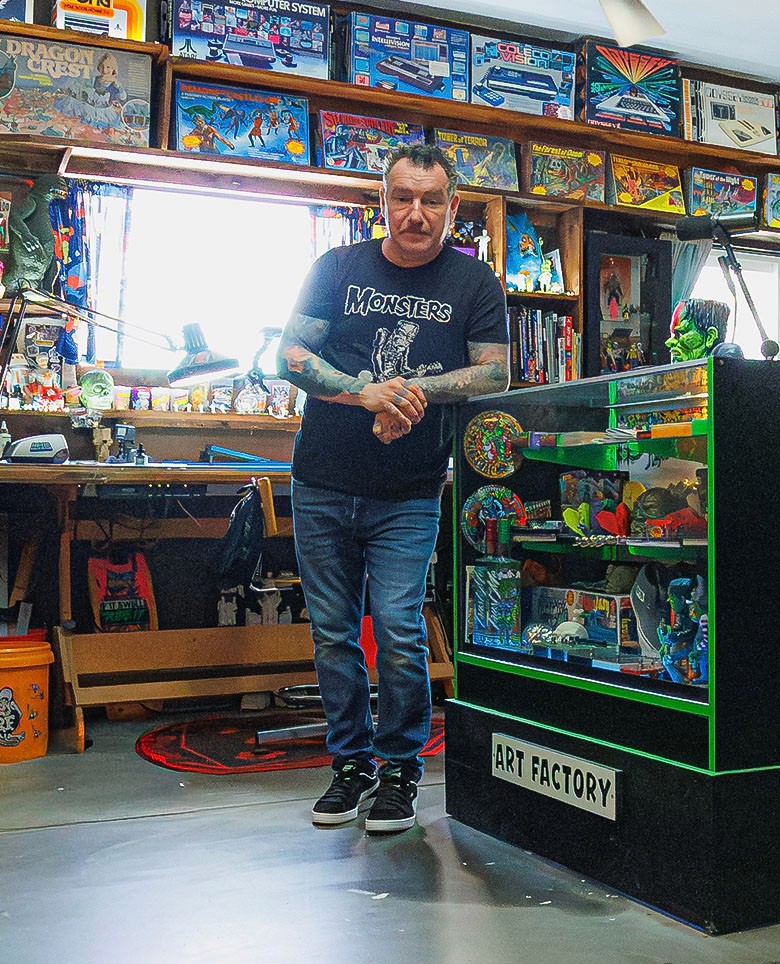 Artist Dirty Donny Gillies standing in his brightly lit studio filled with vintage toys, art supplies, and colorful collectibles, surrounded by shelves packed with models, posters, and pinball machines.