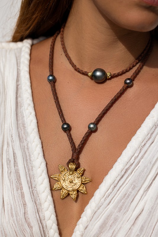 Close-up of a woman wearing Manos de Oro Saint Barth handcrafted jewelry, featuring a radiant gold sun pendant and black pearls on braided leather, capturing island elegance and artisanal soul.