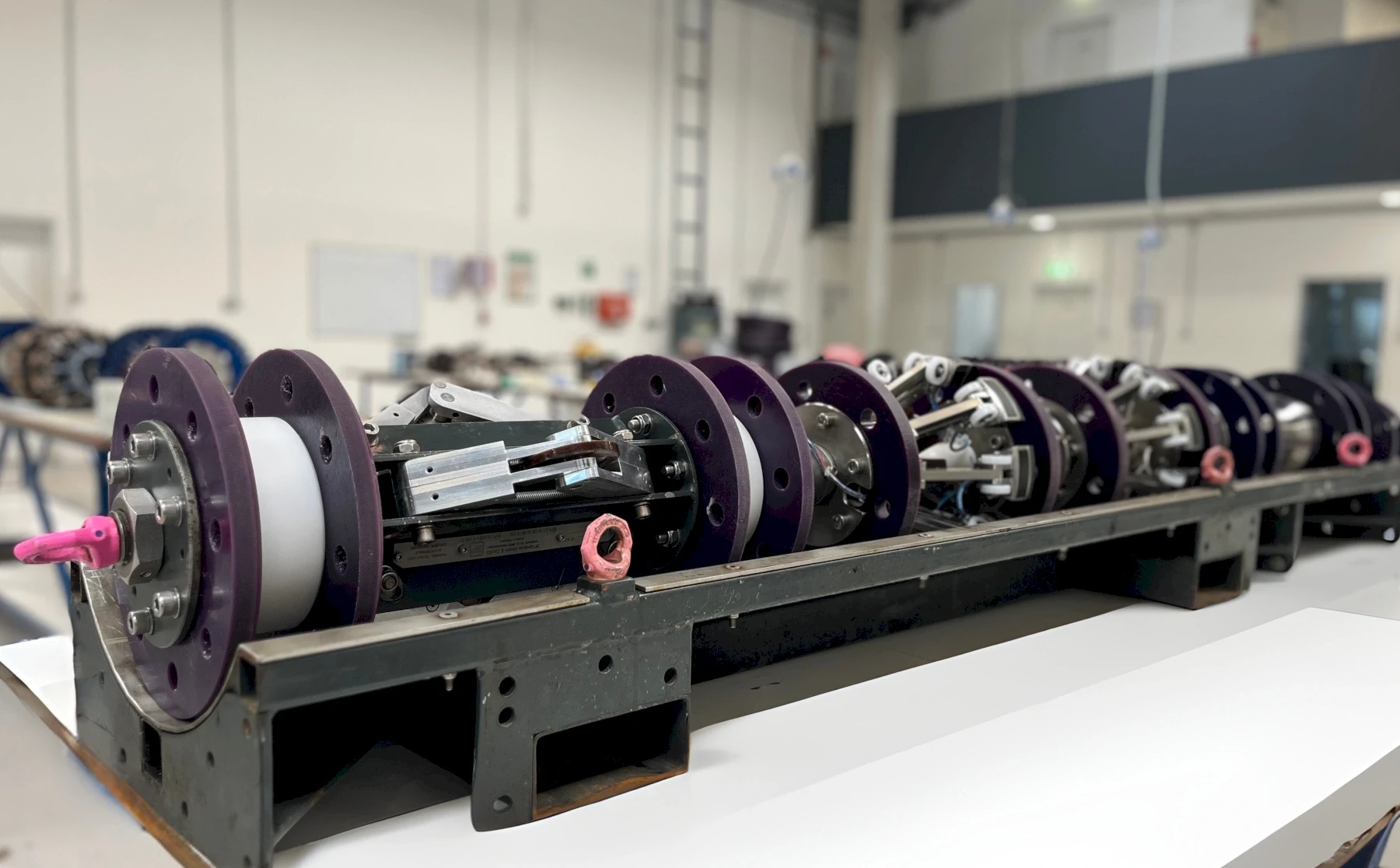 Line of mechanical parts with purple flanges, white cylinders, and various metallic components on a workbench.