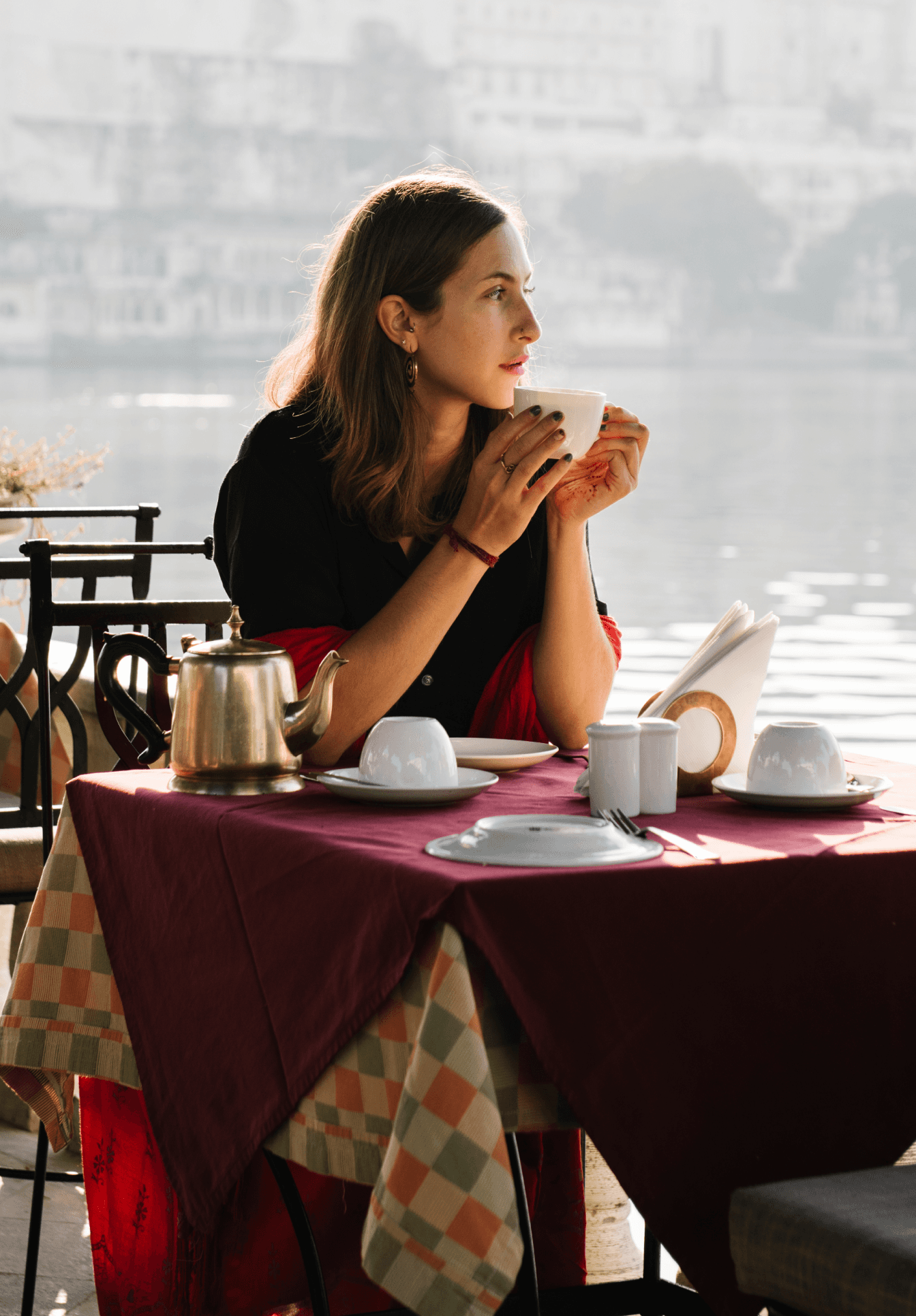 Una mujer sentada en una mesa al aire libre junto al agua, disfrutando de una bebida caliente. La escena es tranquila, con una tetera y tazas en la mesa, lo que sugiere que está relajándose en un café con una hermosa vista al lago.