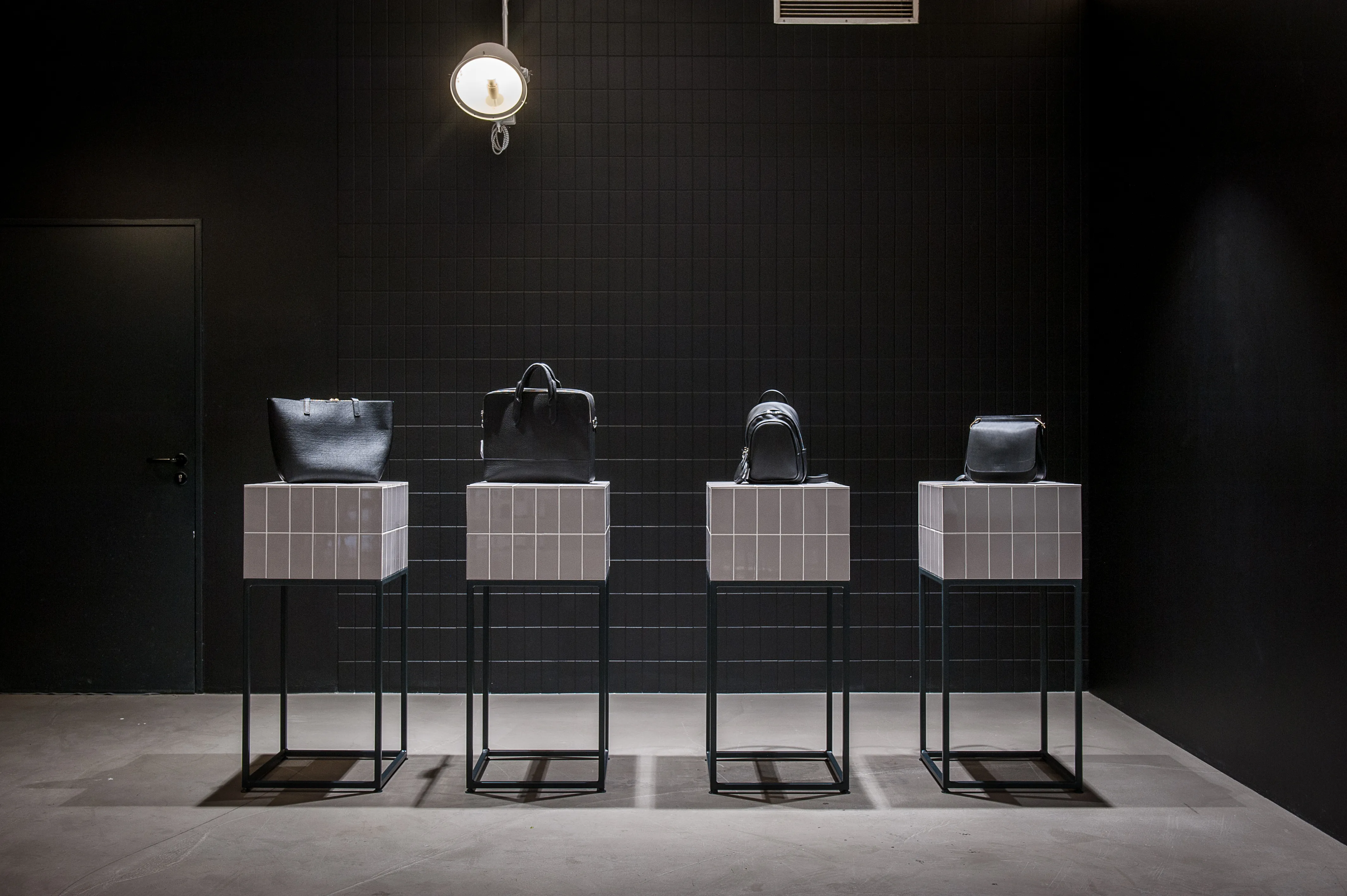 Four black leather bags presented on white tiled pedestal columns with black steel bases in a dark room with black tiled wall and a single spotlight at the 17 FASHION BRANDS showroom.