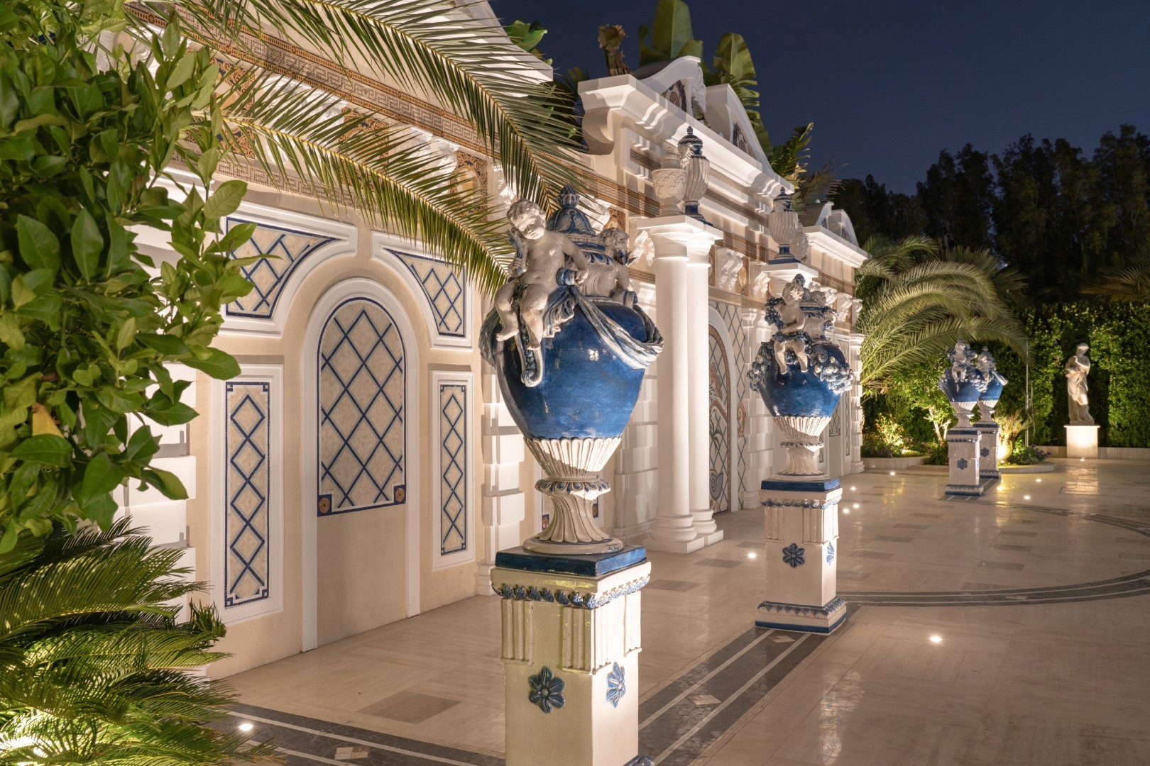 The image showcases an elegant, Mediterranean-style courtyard at night, featuring intricate blue and white architectural elements, large ornamental vases on pedestals, lush green palm trees, and a well-lit pathway leading to an ornately designed entrance.