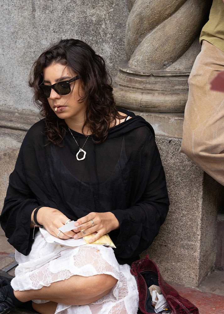 Woman sitting against a stone wall wearing sunglasses and a black top, wearing silver jewelry.