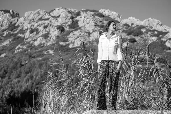 Céline ESCAND, femme élégante se détendant au bord d’un lac au coucher du soleil, portant un manteau marron, ambiance naturelle et apaisante – Le Baume Intégral.