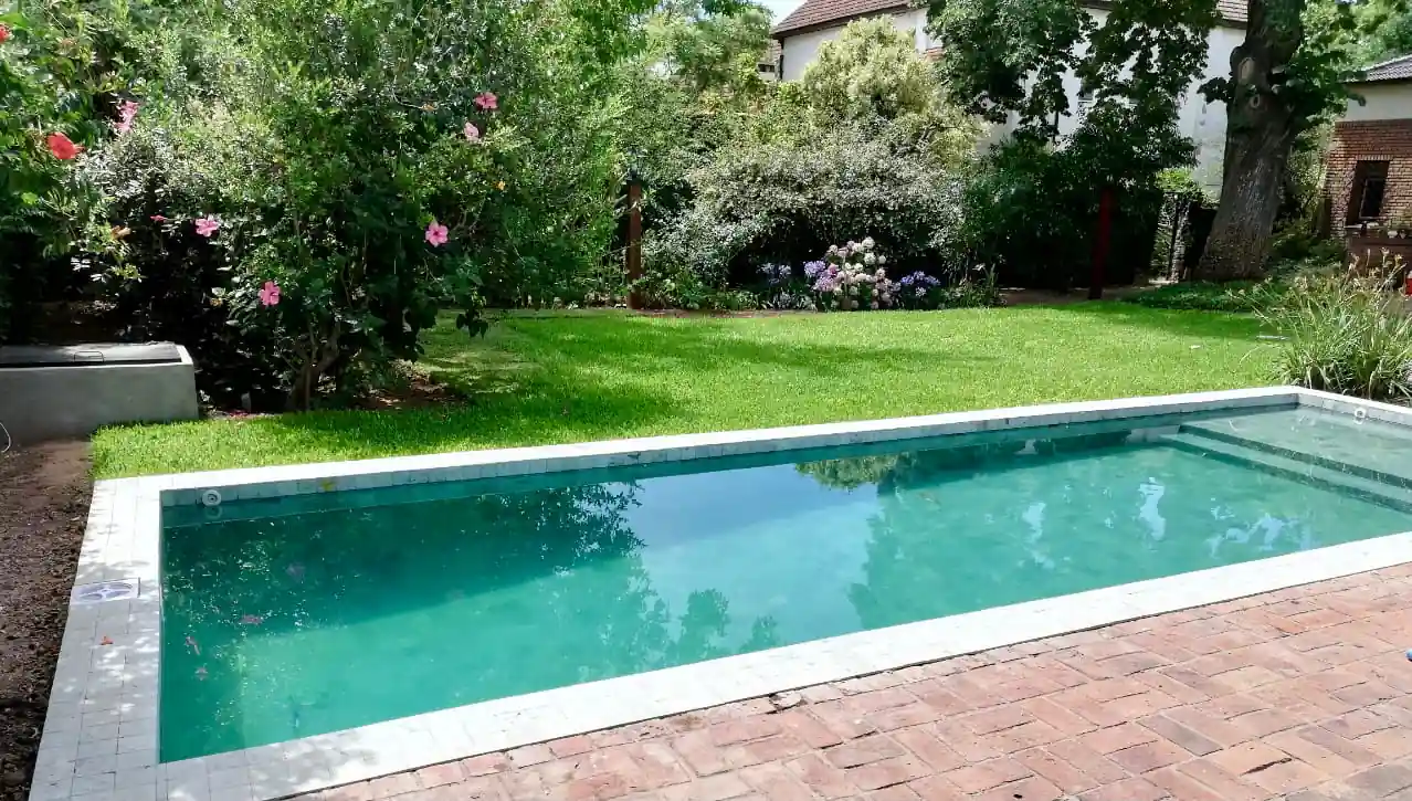 Clásica pileta rectangular de color turquesa con solárium rústico de ladrillos, rodeada de un amplio jardín con flores y árboles.