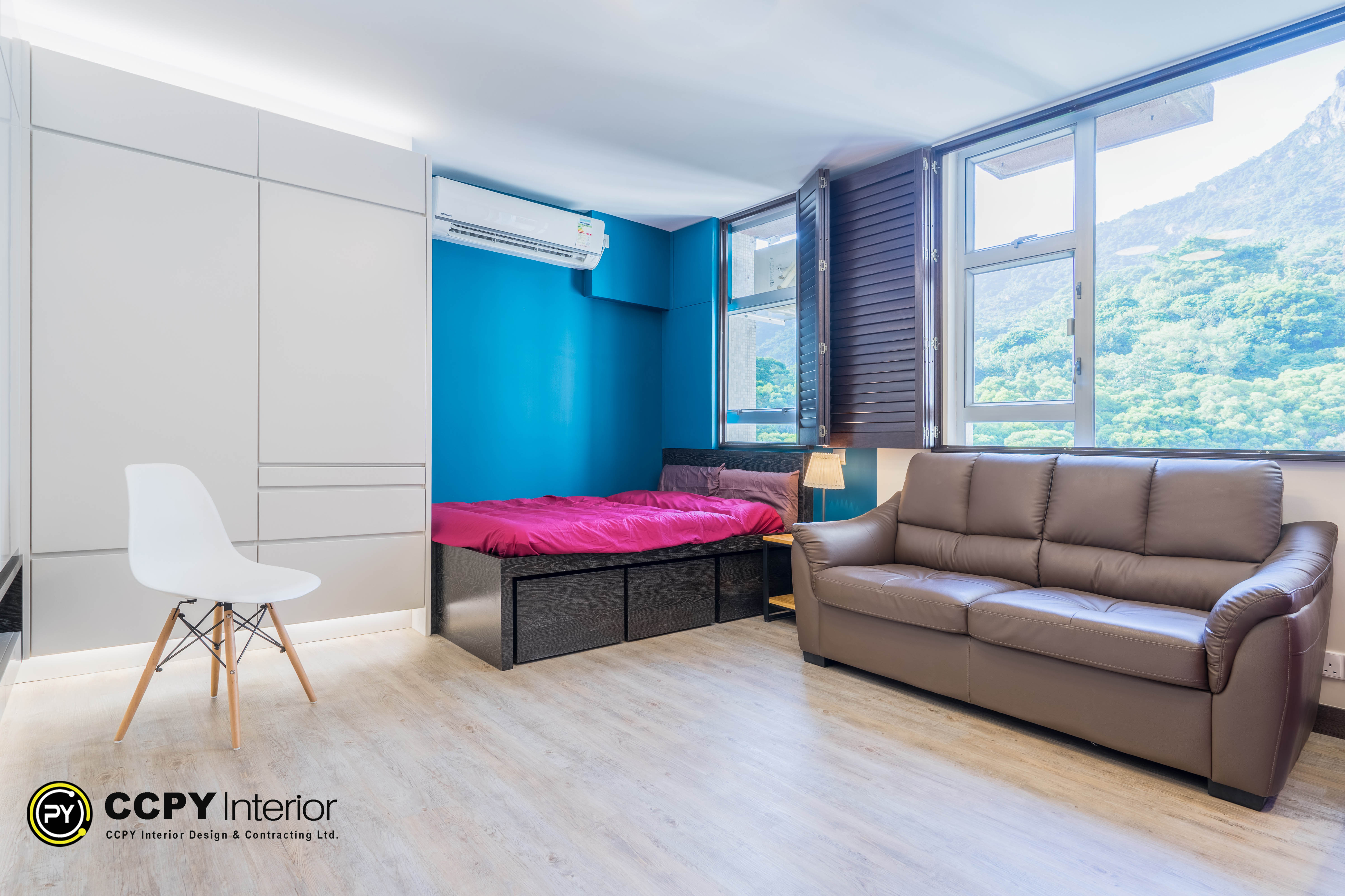 Key Design ElementsSpace-Saving Platform Bed: A dark-toned custom platform system that functions as a sleeping area while integrating deep under-bed storage drawers to reduce visual clutter.Bold Color Blocking: A vibrant teal/deep blue accent wall provides a striking backdrop that adds depth and personality to the compact space.Comfortable Living Set: A brown leather sofa positioned directly alongside the windows, optimized to receive natural light filtered through dark wood louver window shutters.