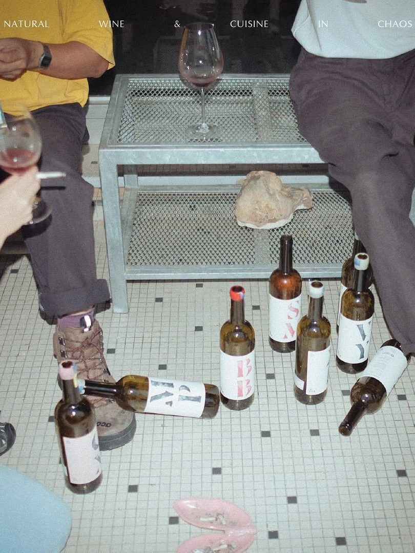 A gathering scene with people seated around a coffee table, surrounded by multiple empty wine bottles and a couple of wine glasses. The floor features a tiled pattern, and a shell is placed on the table.