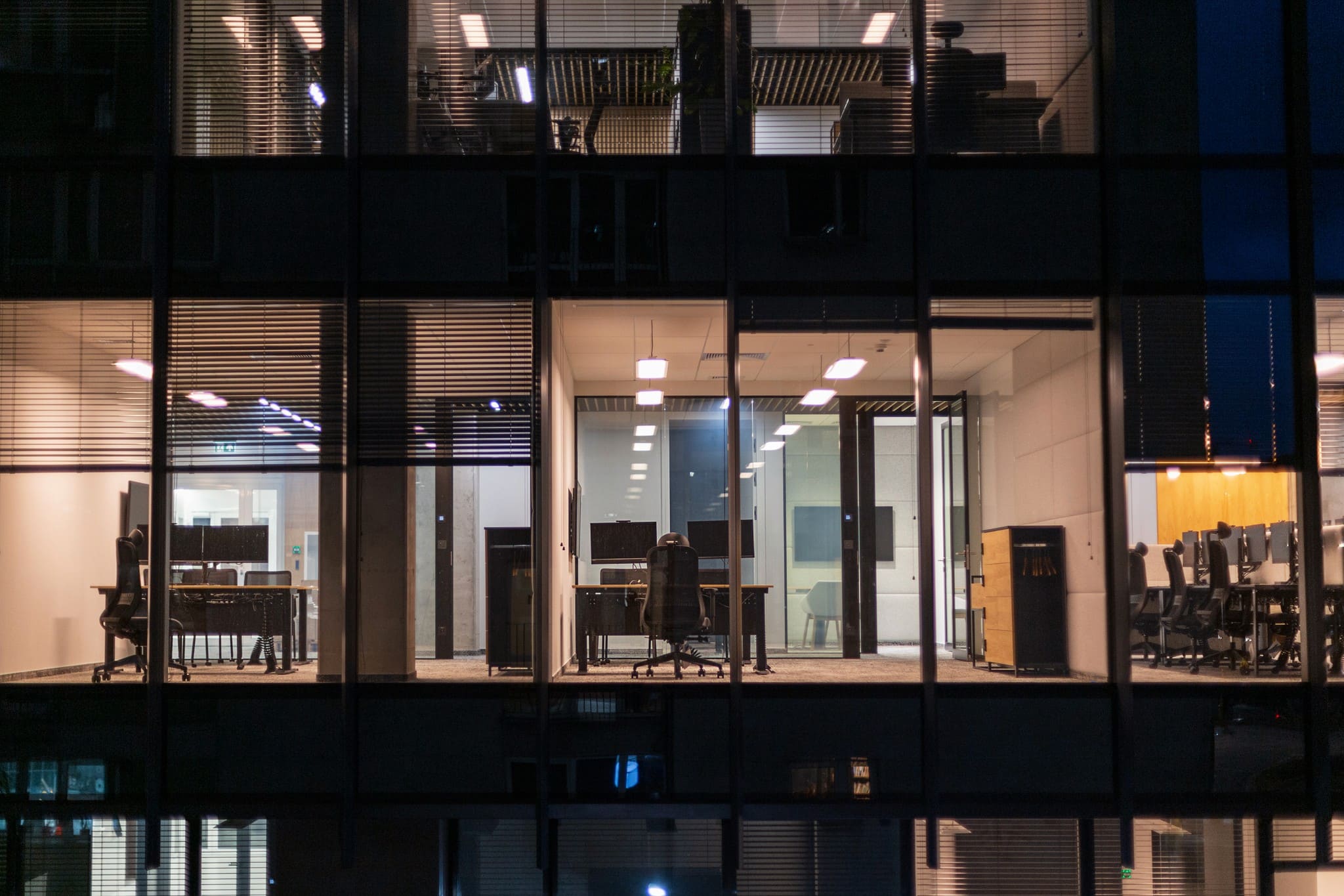 An exterior nighttime view looking into illuminated, empty office spaces through the large windows of a modern building facade.