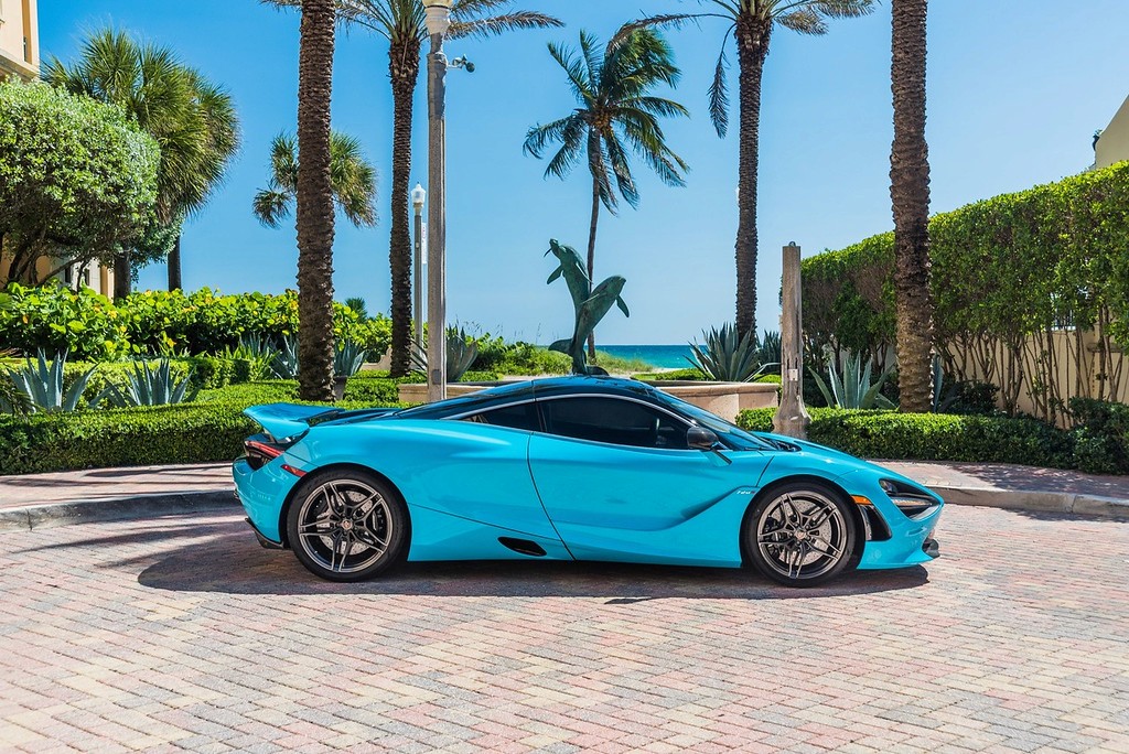 A stunning blue McLaren 720S with dihedral doors open, parked in a luxurious setting with palm trees and an elegant building in the background. This exotic supercar features a sleek design, powerful twin-turbo V8 engine, and unparalleled performance, making it the perfect choice for a luxury car rental experience.