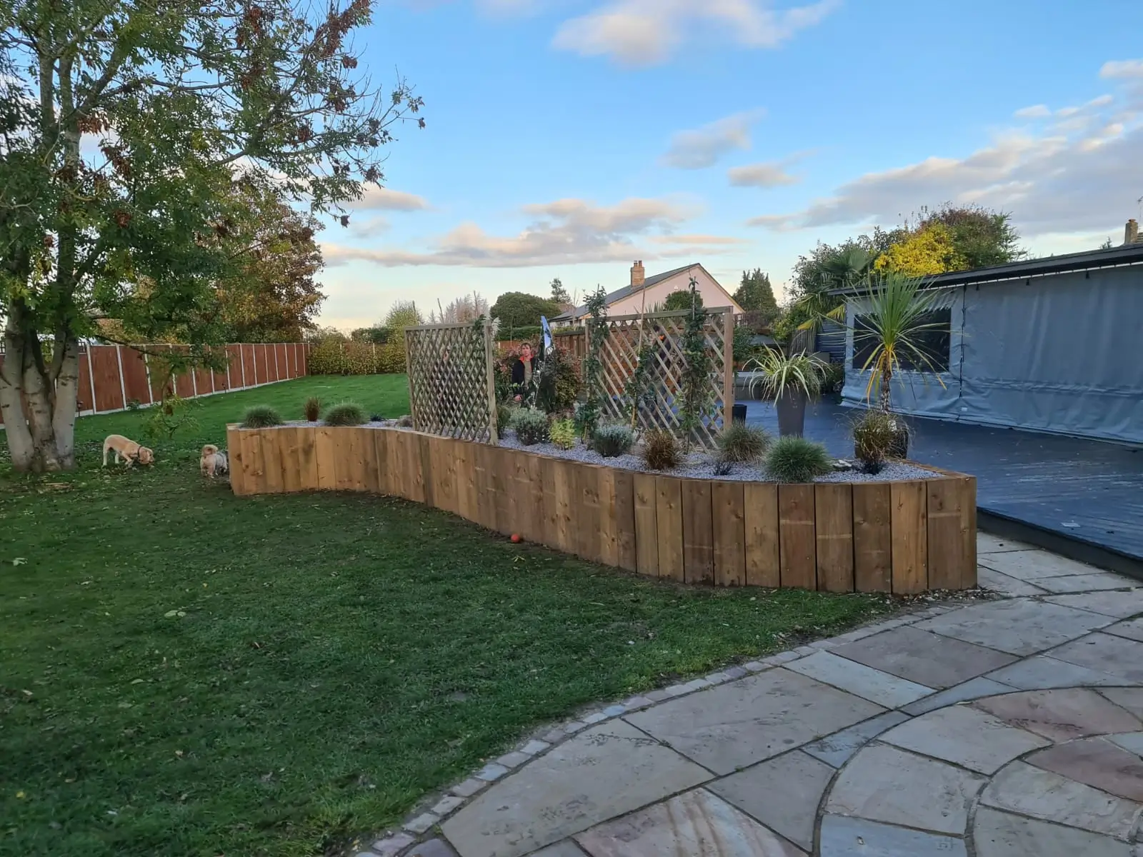 A garden scene with wooden raised beds, greenery, and a pathway under a blue sky with scattered clouds.