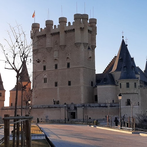 A stone castle with multiple towers and a flag atop one. Leafless trees and a paved path in the foreground, with a few people walking.