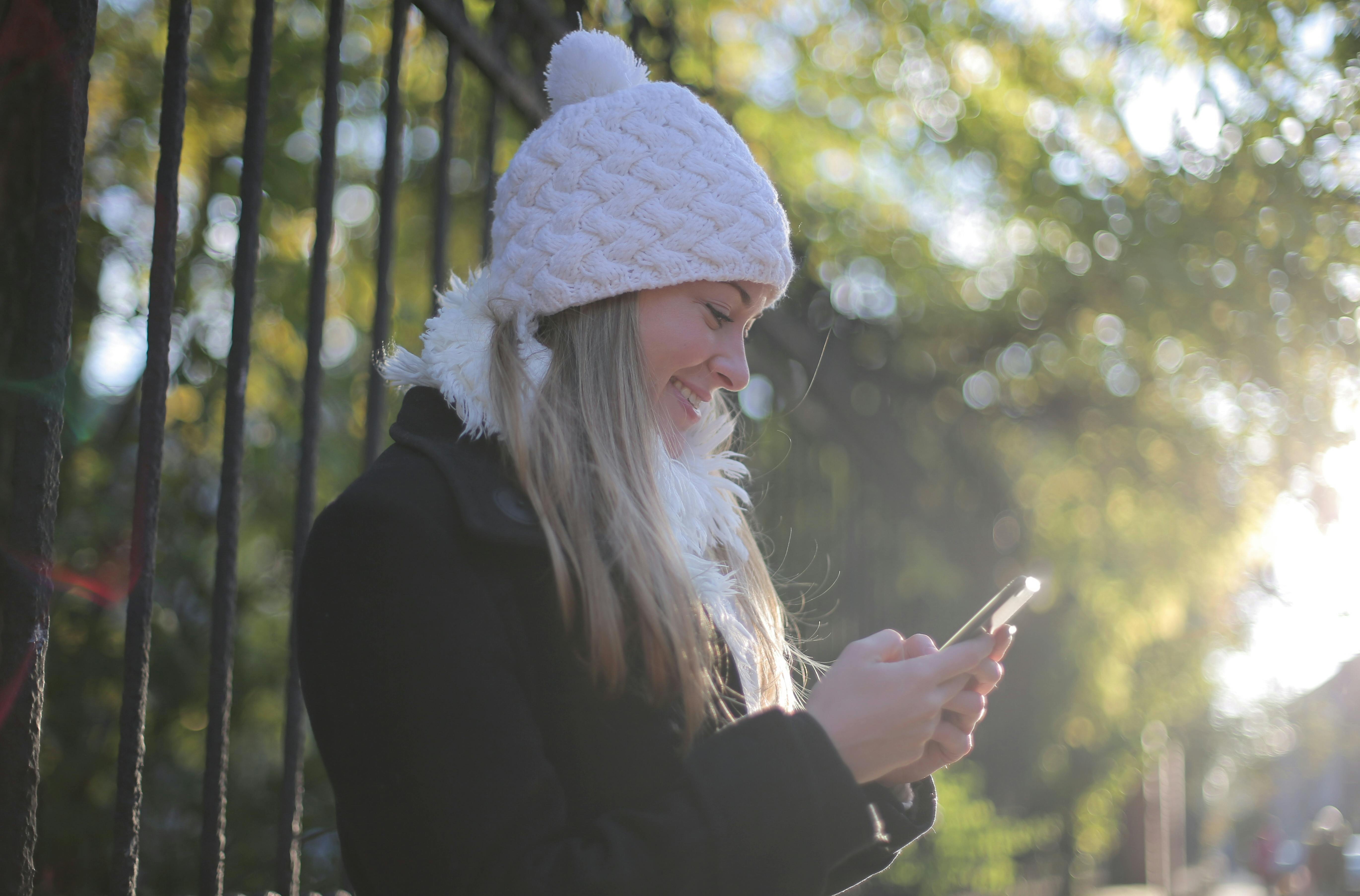 A smiling woman in a white winter hat looking at her smartphone outdoors, representing a Canadian resident finding relief from the cost of living by reading our weekly financial deals newsletter.