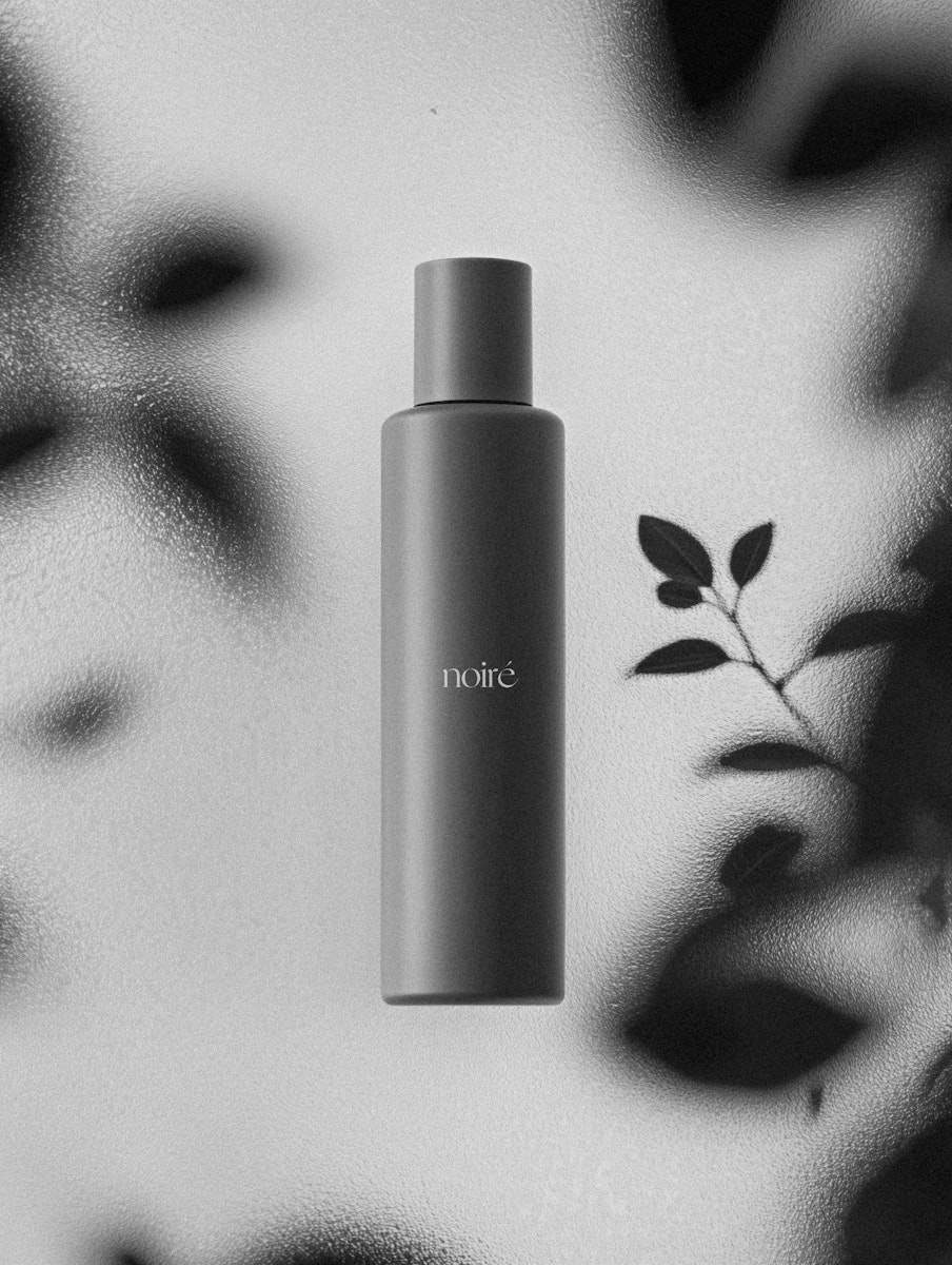Minimalist black bottle with 'noiré' branding, set against a textured, frosted background with abstract shadows and leaf silhouettes.