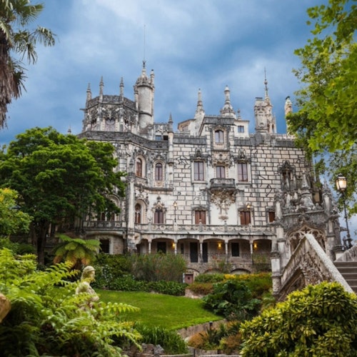 An ornate historic building surrounded by lush greenery and stairs, with a cloudy sky overhead.