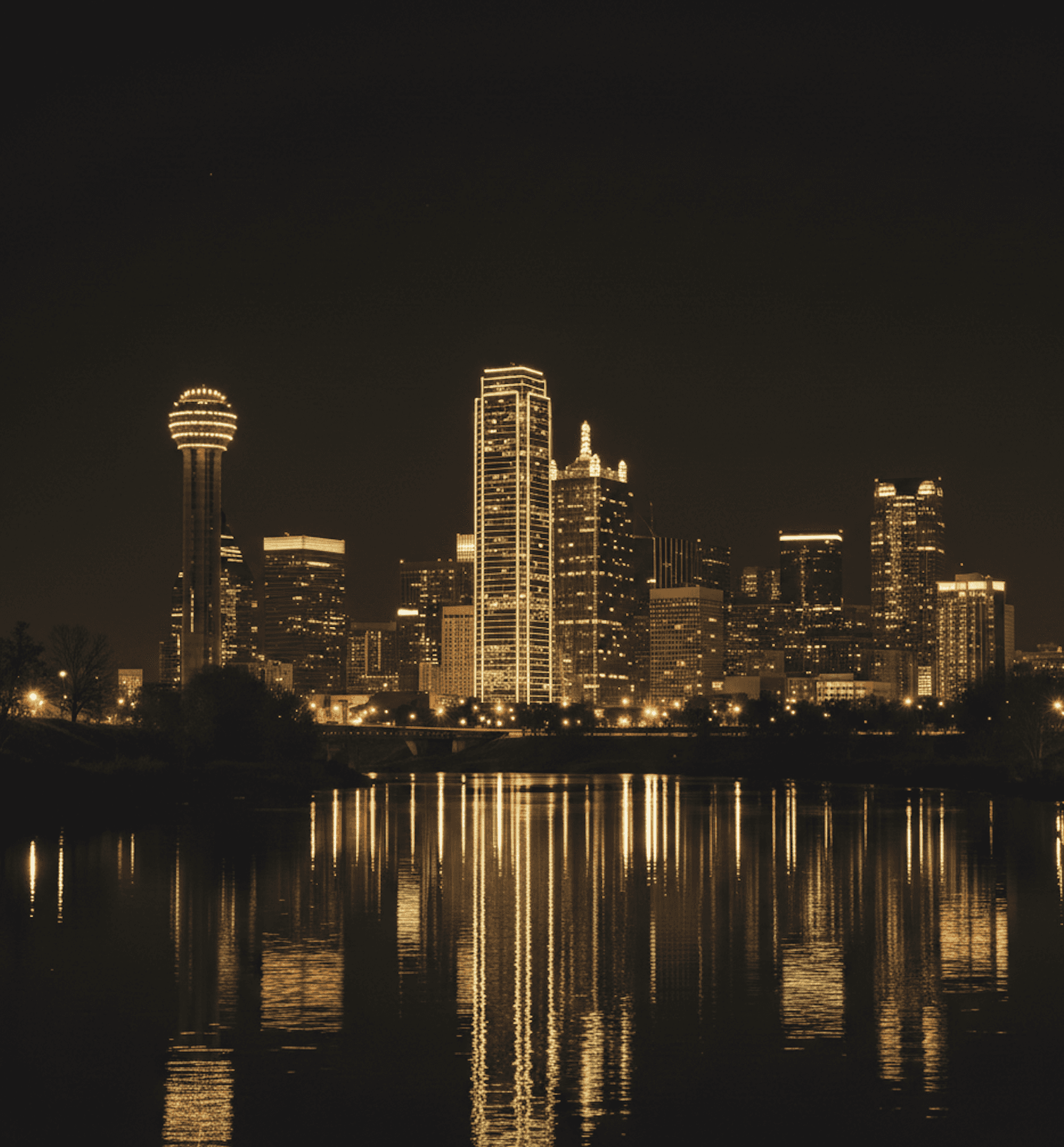 The Dallas city skyline illuminated at night with golden lights reflecting on the water in the foreground.