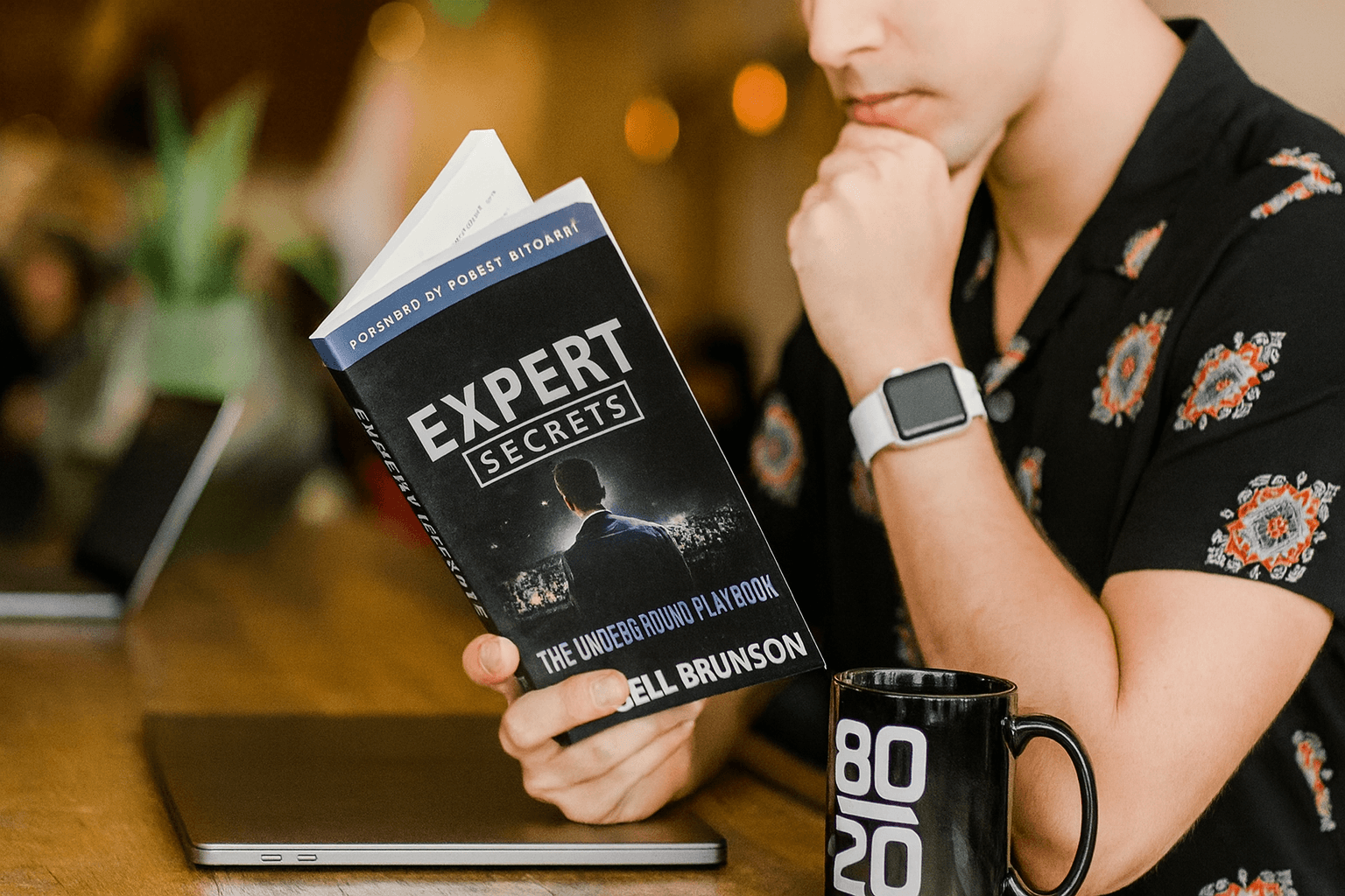 Un homme lisant un livre intitulé "Expert Secrets" dans un café, entouré de personnes travaillant sur des ordinateurs, illustrant l'importance de collaborer avec des experts pour optimiser les campagnes Amazon Ads.