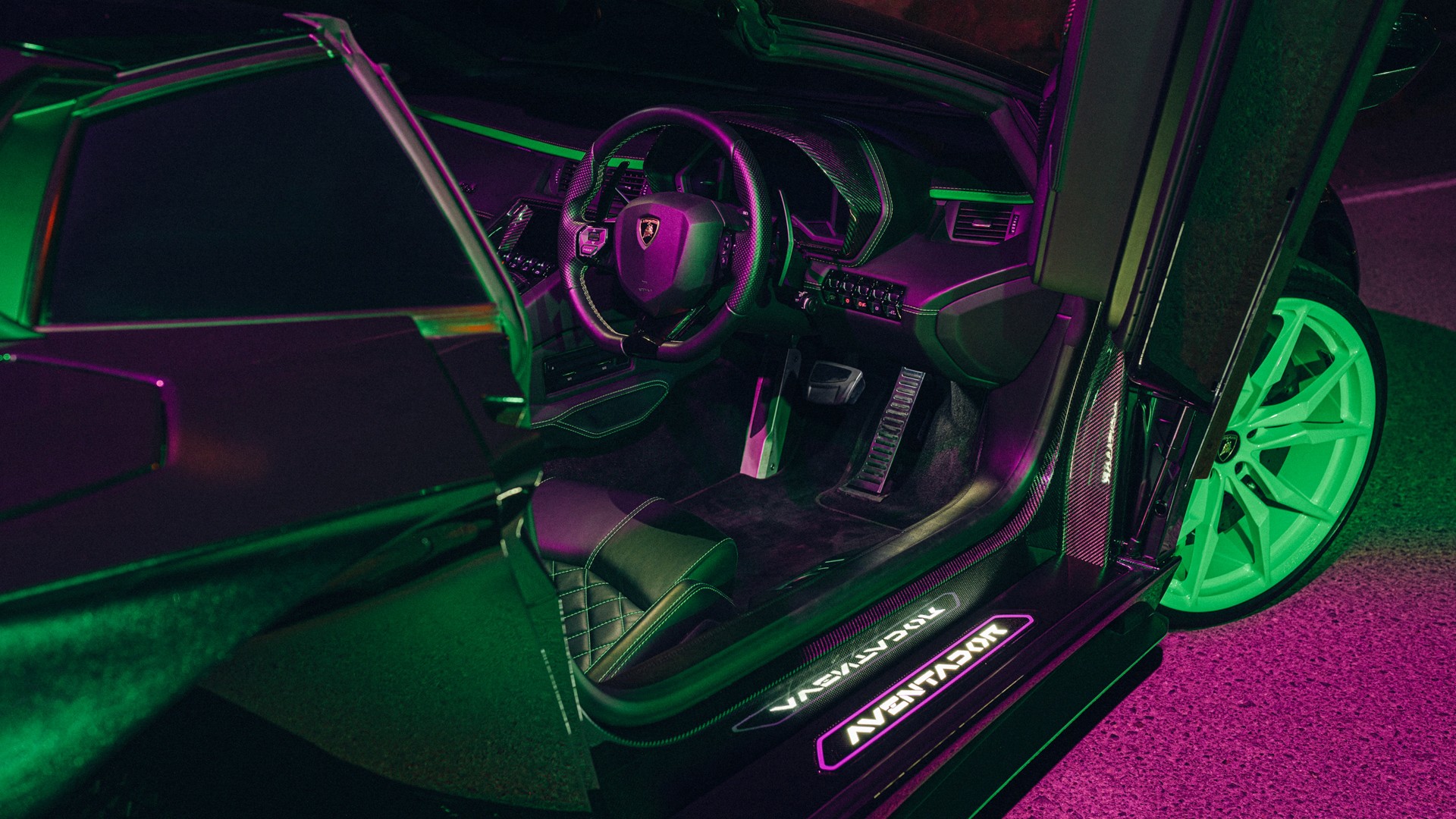 Interior of a Lamborghini Aventador through the raised scissor door, showing the carbon-trim steering wheel, diamond-stitched seats and an illuminated "AVENTADOR" door sill, lit in green and magenta neon with a green wheel in frame.