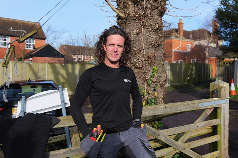 A person with long hair stands by a wooden gate, smiling against a backdrop of houses and blue sky.