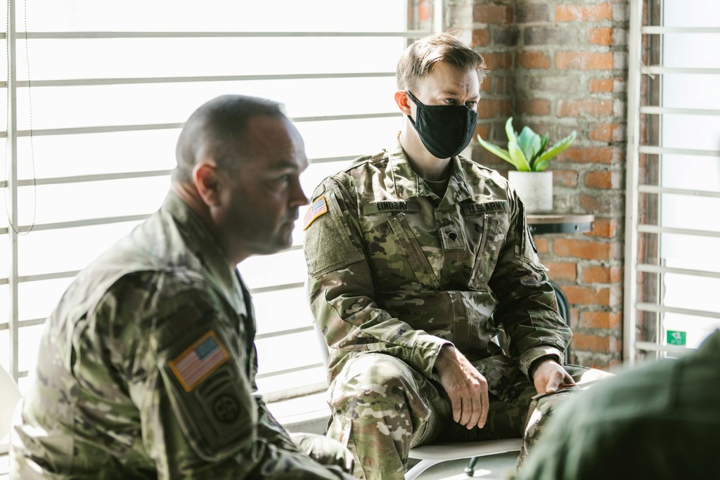 The image shows two military personnel in camouflage uniforms and face masks seated indoors, engaged in a conversation. They are in a well-lit room with natural light coming through horizontal blinds, and a small potted plant on a shelf against a brick wall adds a touch of greenery. The scene conveys a sense of professionalism and attentiveness, highlighting the soldiers' dedication and camaraderie in a relaxed setting.