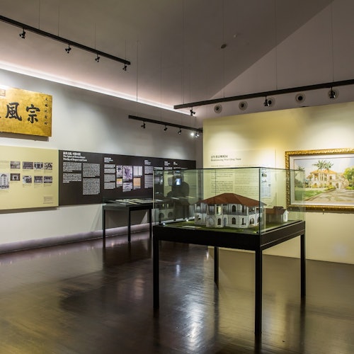 A museum exhibit featuring historical displays on walls, a glass case with a building model, and framed artwork on the right.