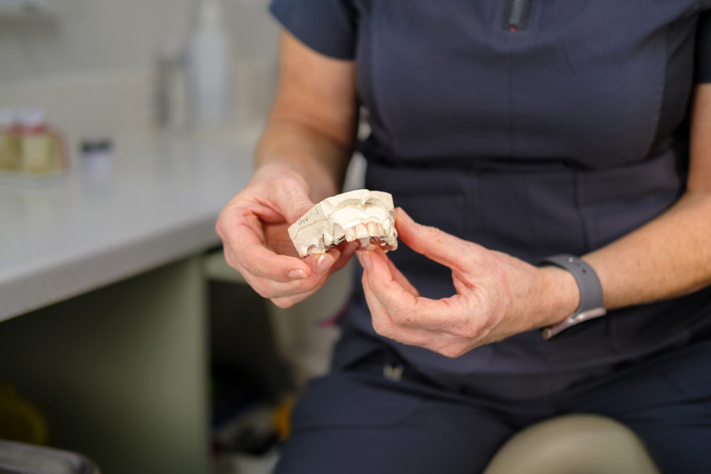 A person in a dark uniform holds a dental mold in a clinical setting, with a desk and dental tools visible in the background, highlighting dental care and orthodontic practices.