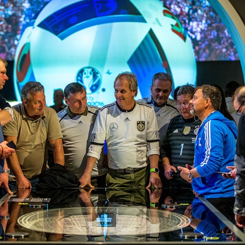 一群男人，有些穿著運動衫，圍在一個互動展示前，背景是一個大型足球和體育場的場景。