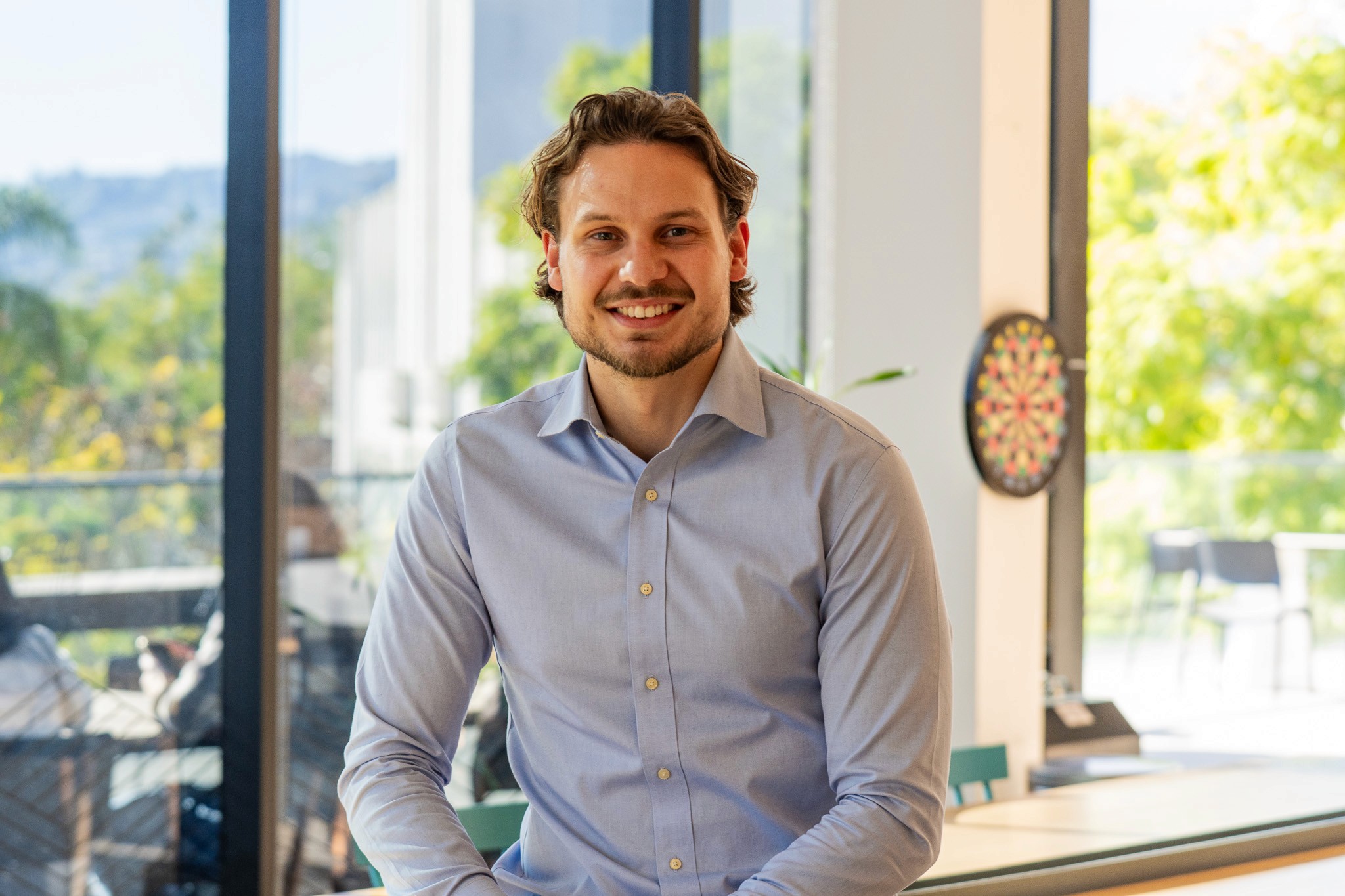 Smiling young woman with long hair standing against a dark green background, holding a finger to her chin.