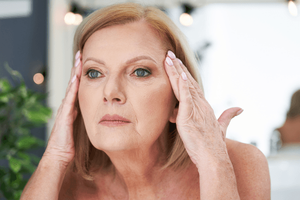 An image of a woman looking at herself and pulling on her face to make her wrinkles go away with a plant and white in the background