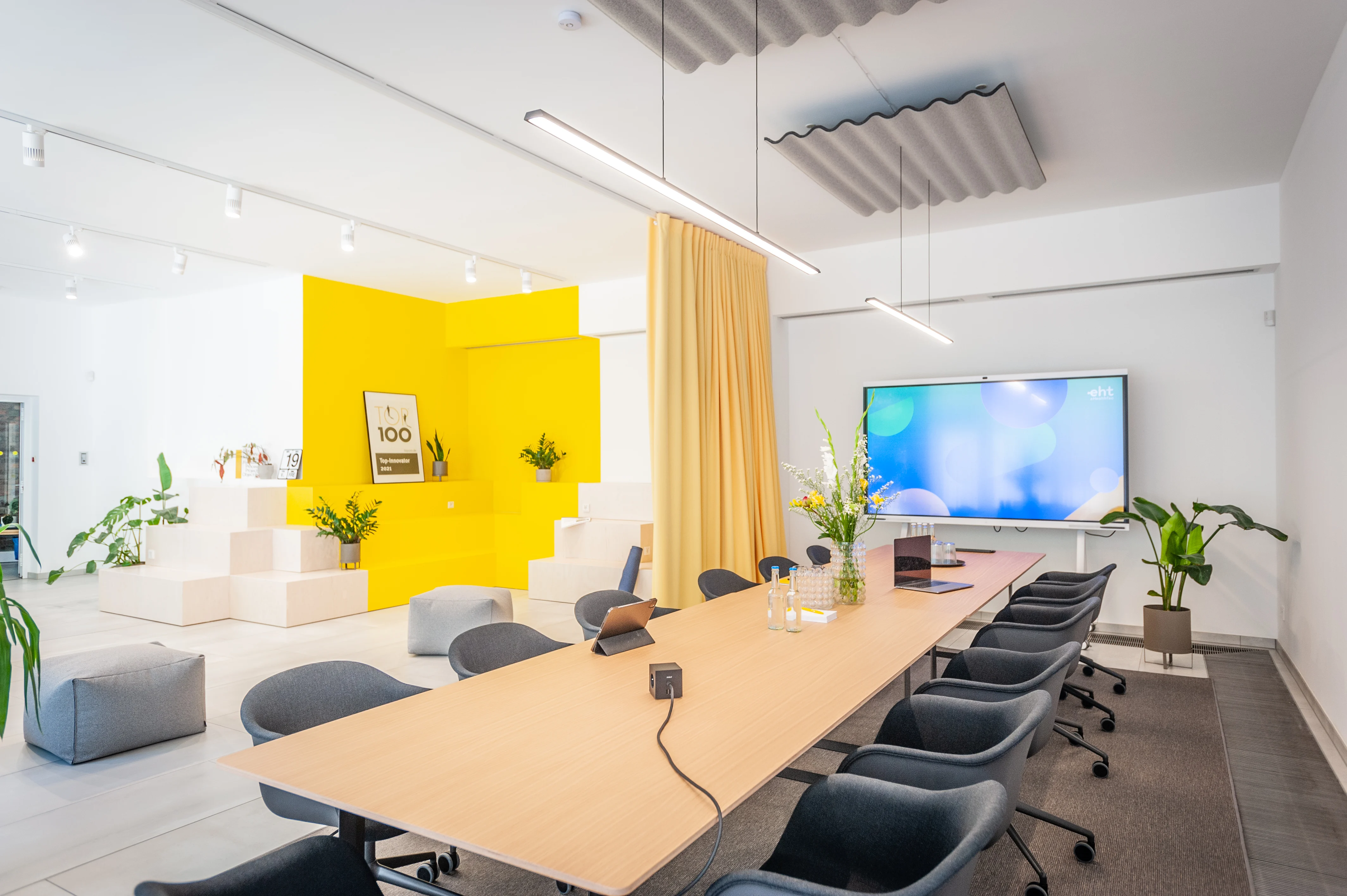 Large conference room at EHEALTH-TEC with a long oak table, grey upholstered swivel chairs, a wall-mounted video screen, corrugated acoustic ceiling panel, yellow curtain and stepped yellow-and-white seating corner.
