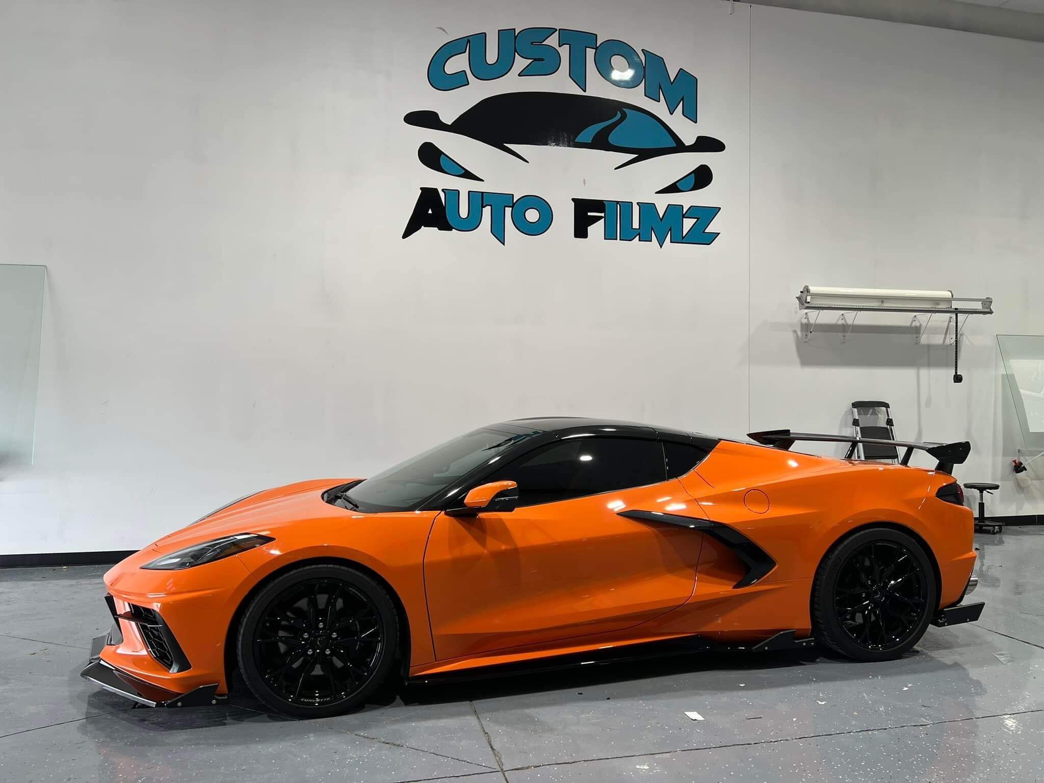 A bright orange Chevrolet Corvette C8 with black accents is parked indoors on a polished gray floor.