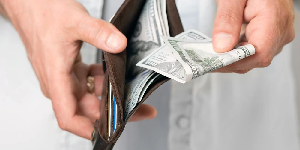 A man in casual clothes paying his bills holds a brown wallet with hundred dollar bills in his hand.