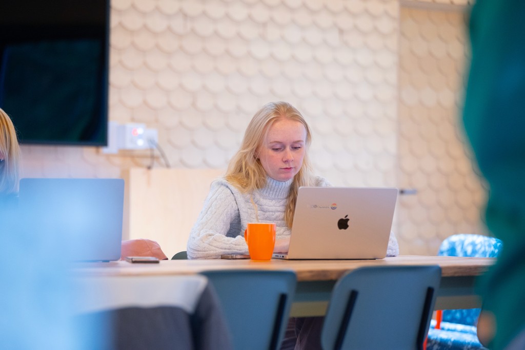 A person sits at a large table in a modern workspace, using an open laptop with an orange mug beside it. Another laptop is visible to the side, and the background features a textured wall and soft, warm lighting.