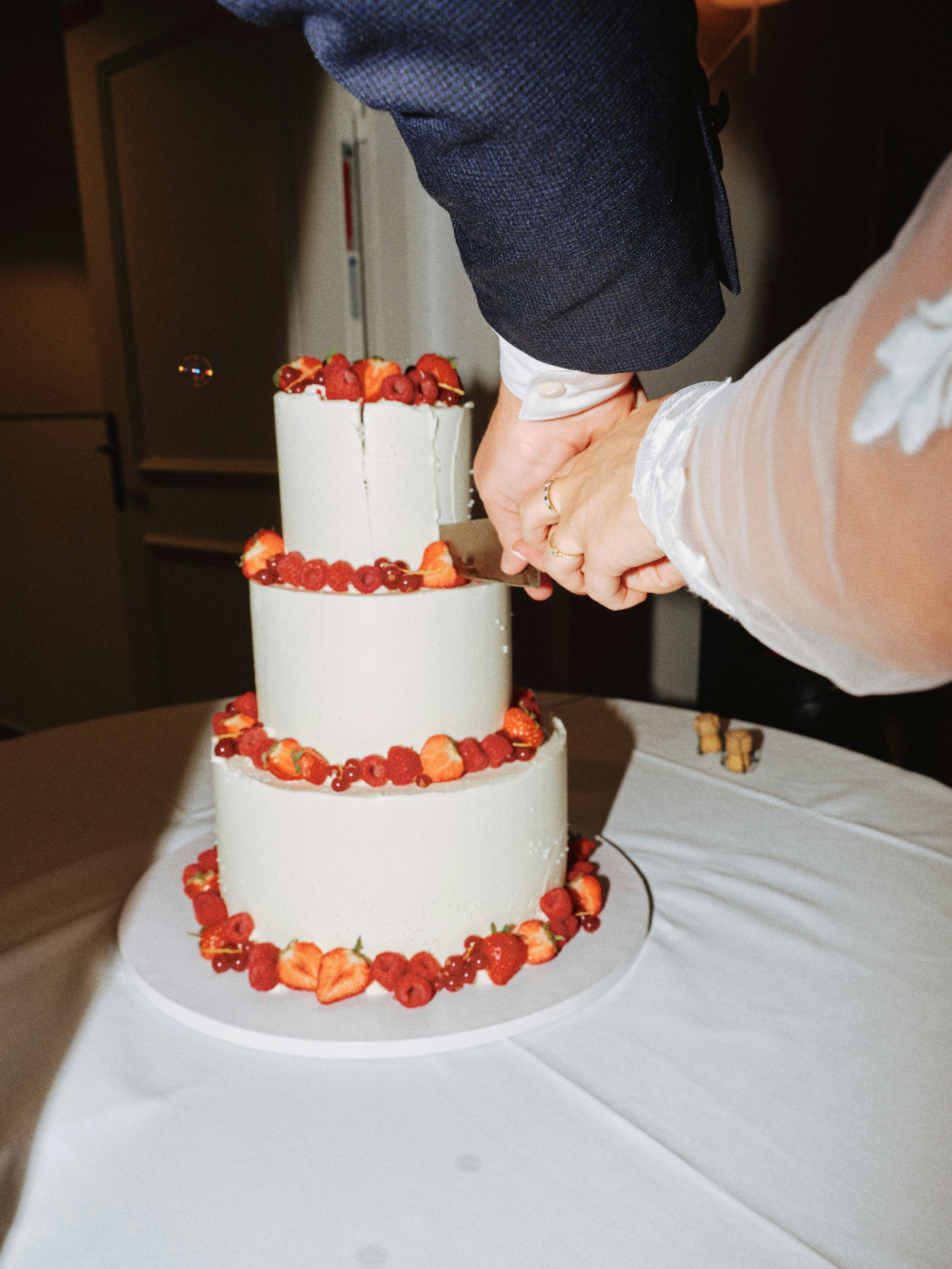 Wedding cake cutting moment with the couple's hands interlocked over the knife, three-tier white cake with strawberry and raspberry trim, warm ambient lighting