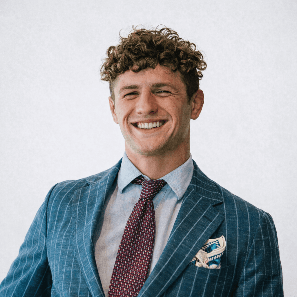 Casual studio portrait of a bearded man with curly hair in a white shirt, arms crossed, on a light gray background.