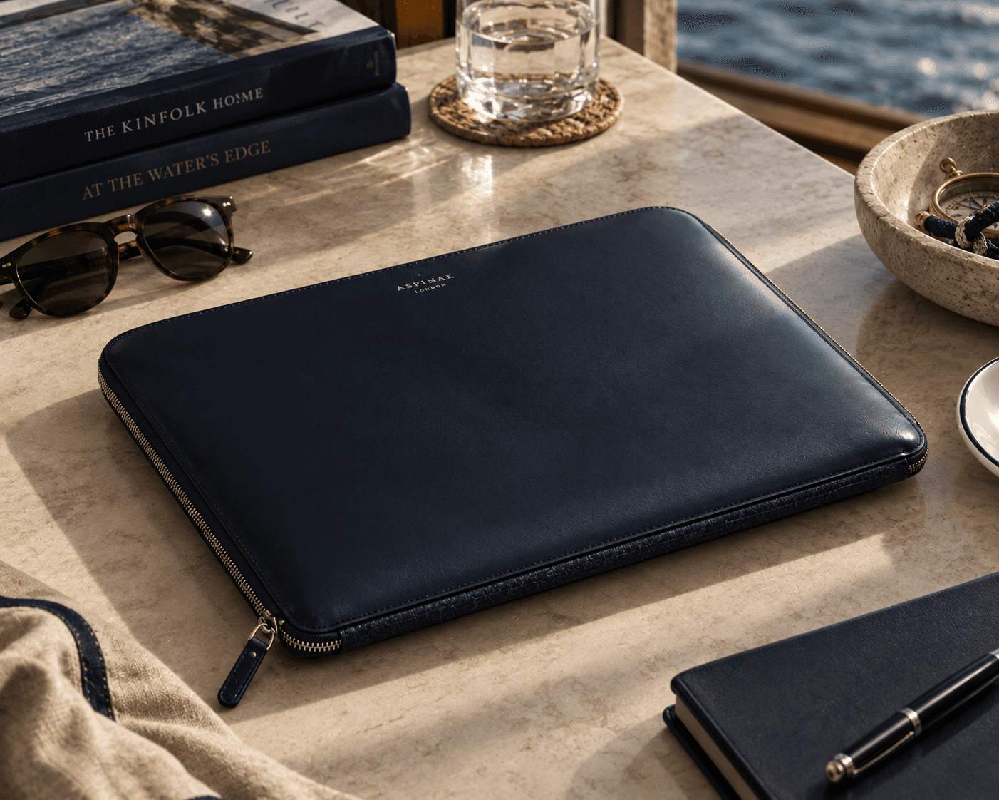 A sunlit luxury accessory flatlay set on a beige travertine stone ledge overlooking the sea. At the center is a dark navy blue leather zip-around laptop sleeve featuring gold embossing that reads "ASPINAL OF LONDON". Surrounding items include stacked coffee table books titled "THE KINFOLK HOME" and "AT THE WATER'S EDGE", tortoiseshell sunglasses, a glass of water on a woven coaster, a small stone bowl with vintage pocket accessories, a navy notebook with a metallic pen, and a folded cream linen towel.