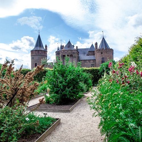 Et stort murstensslot med flere tårne set fra en have fyldt med forskellige planter og blomster under en delvist overskyet himmel.