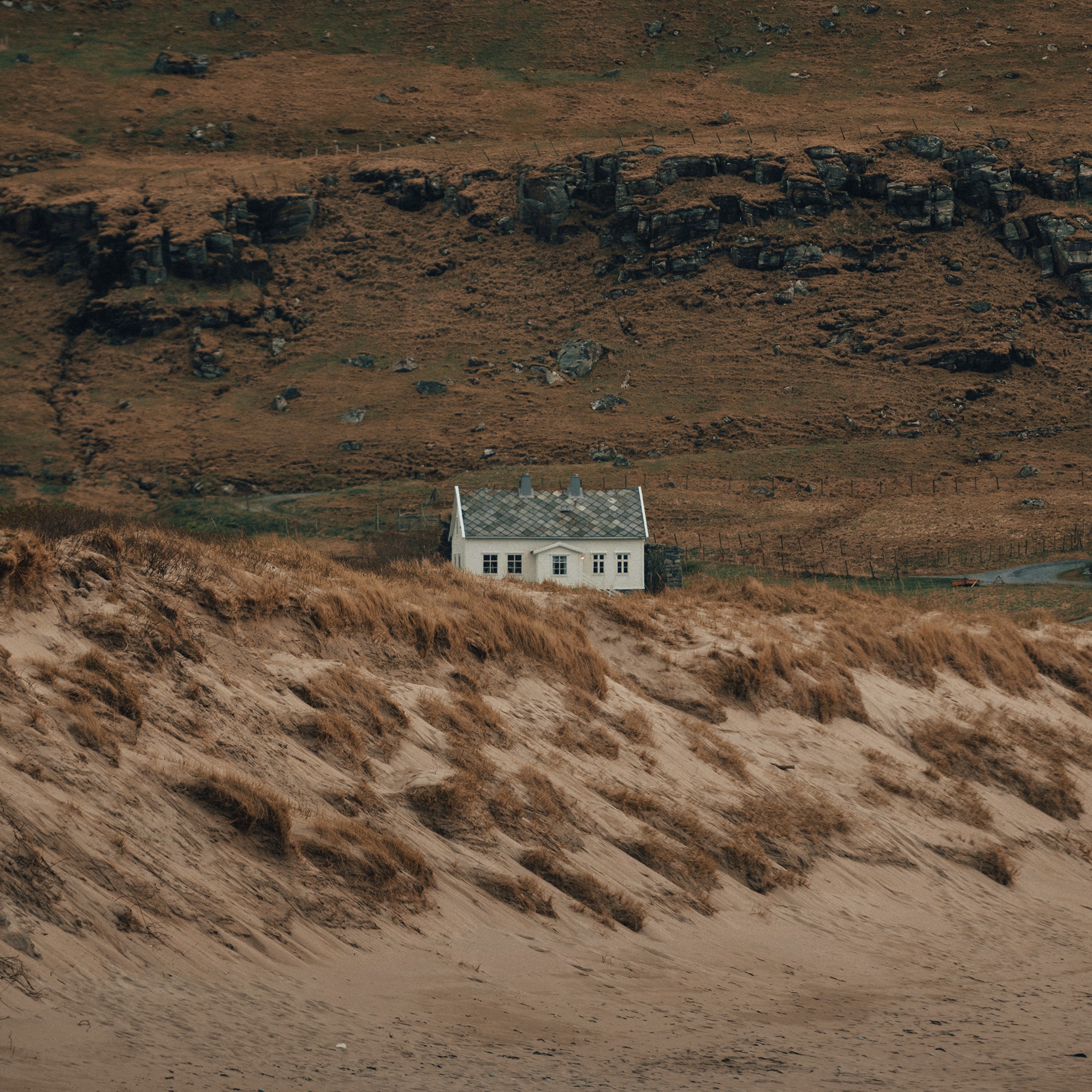 Small white house surrounded by brown grass dunes and rocky hillside in the Faroe Islands