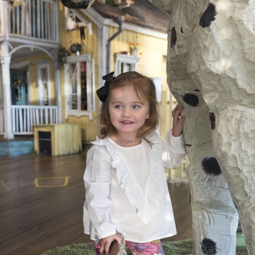 Young girl in a white top stands next to a large, white sculpture with black spots, smiling inside a brightly lit room with wooden floors.