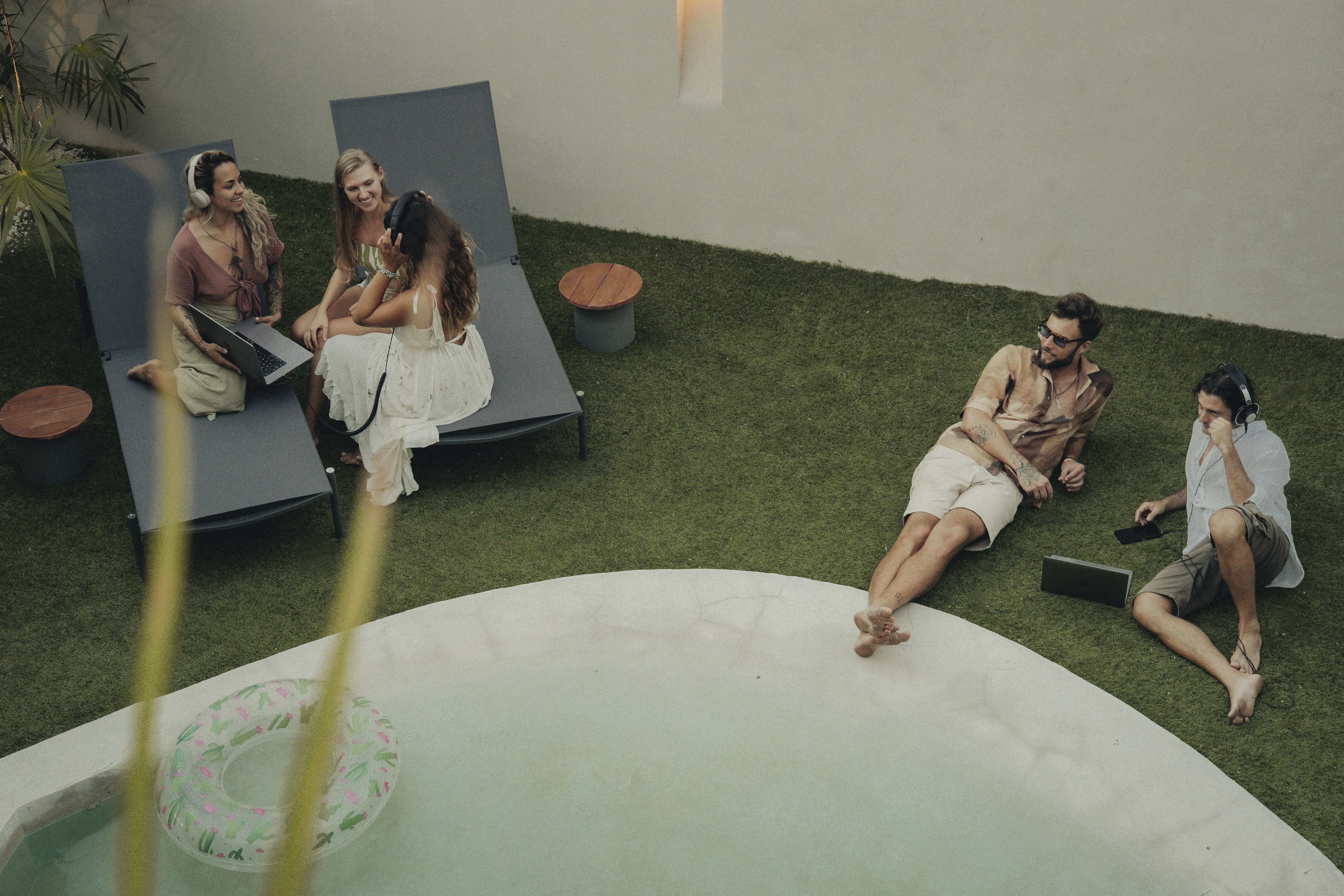 Three women sitting on sunbeds, smiling and talking while looking at a laptop, with two men nearby using a laptop and phone.