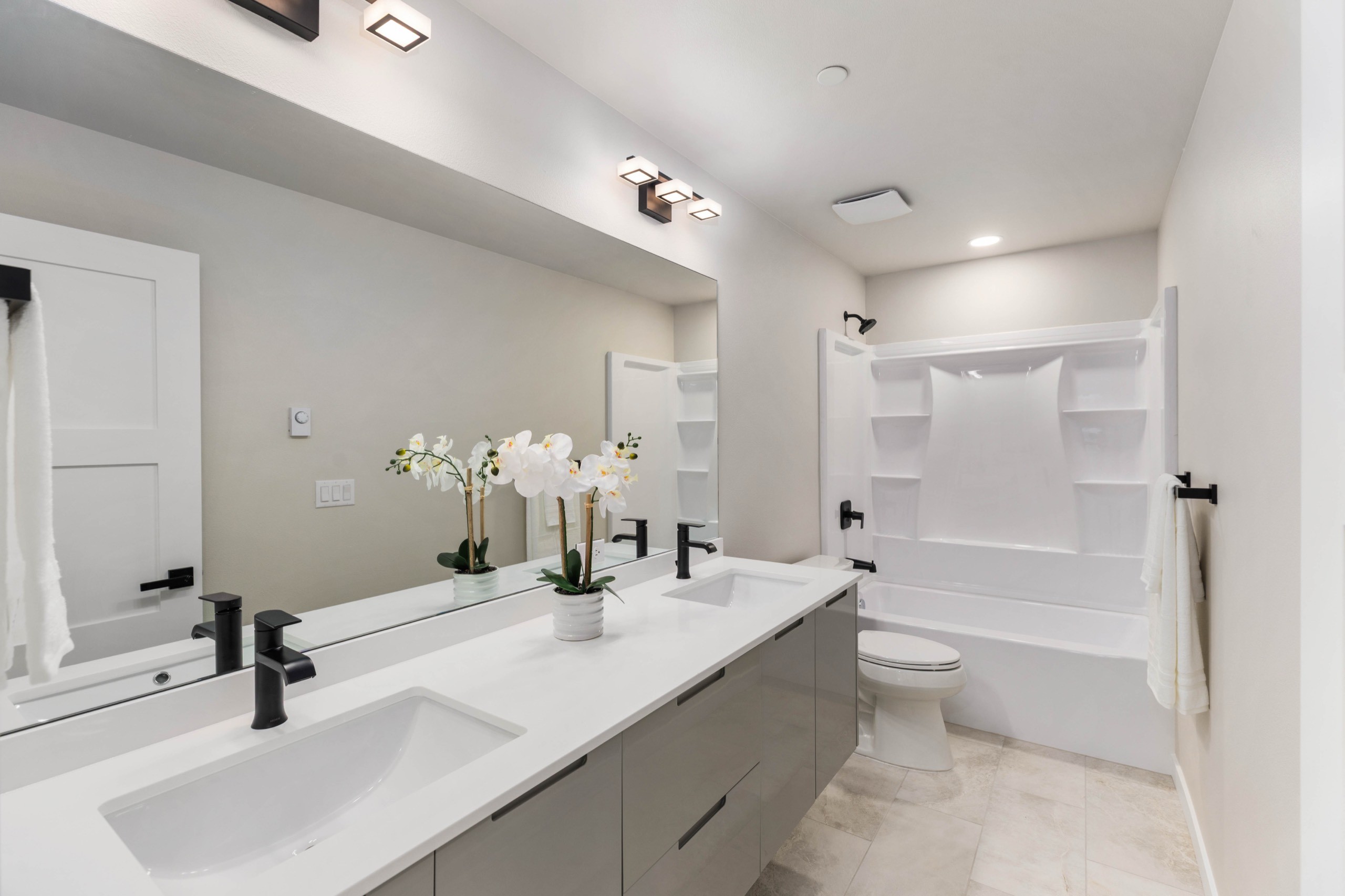 Modern luxury double-sink bathroom with grey floating vanity, white quartz countertop, black fixtures, oversized mirror, and soaker tub with white tile surround