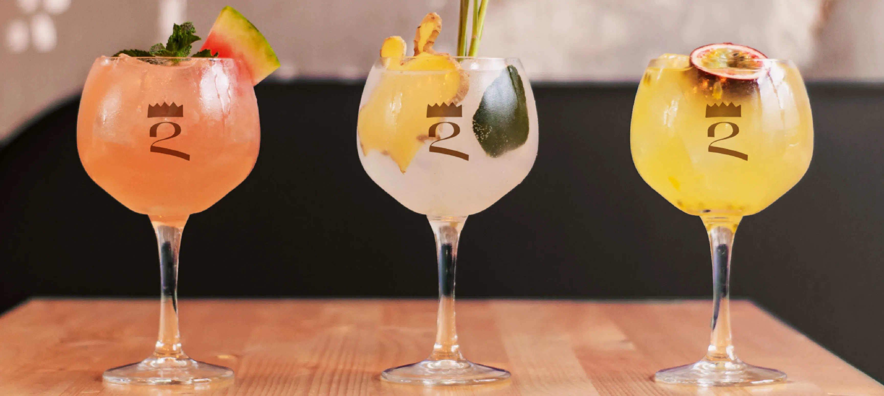 Three fruity cocktails sitting on a wooden table in round wine glasses with a crowned 2 monogram printed in gold on the glasses. 