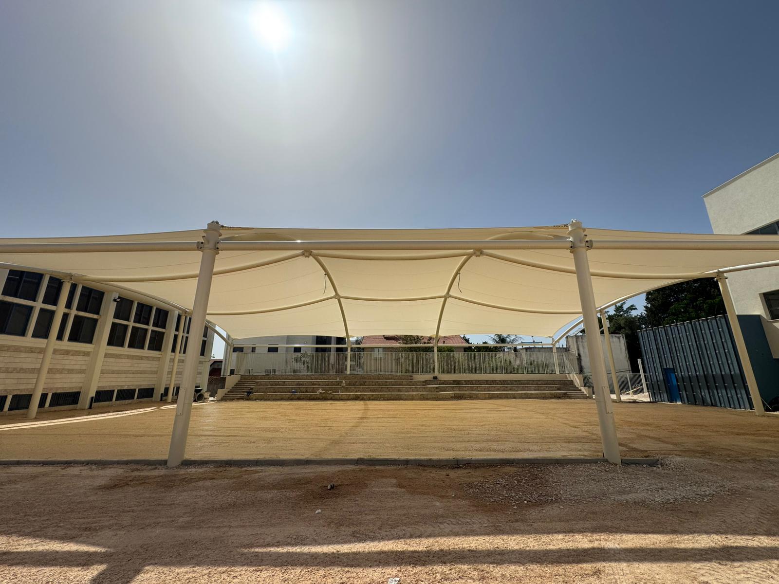 Large beige sunshade sails over an open area with steps and buildings in the background under a sunny sky.