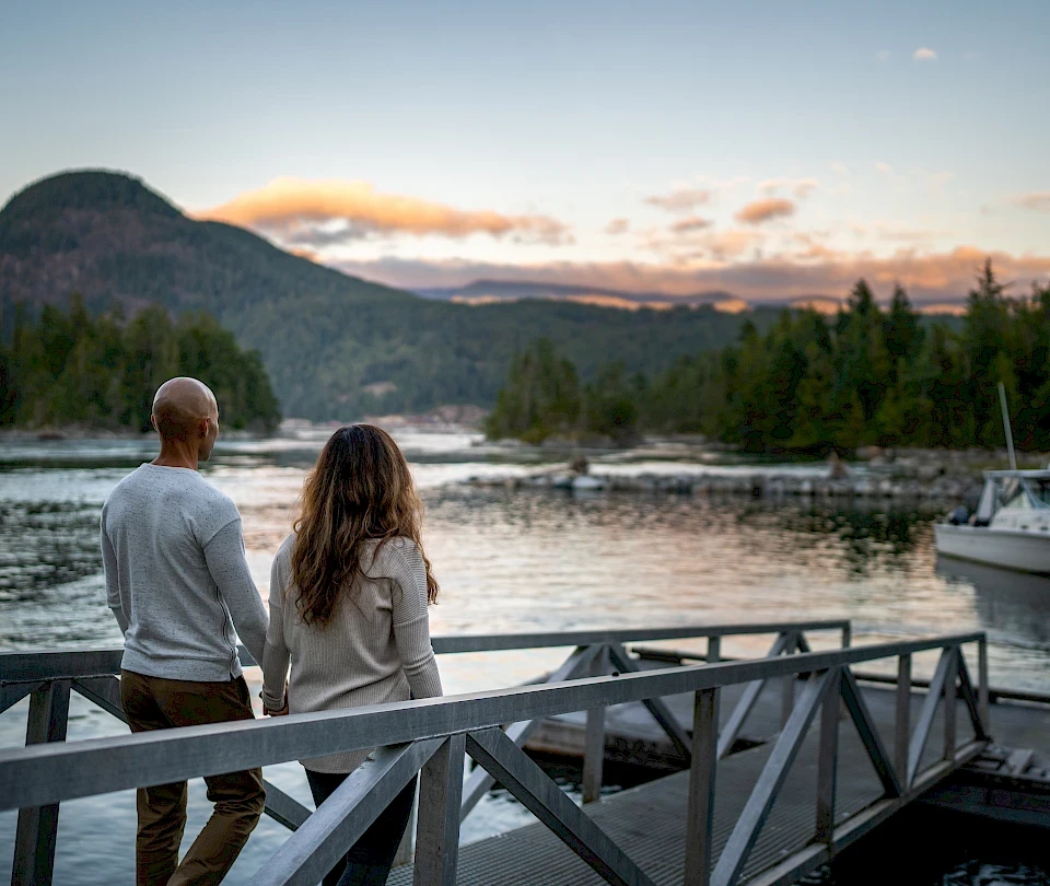 Ein Paar steht Hand in Hand auf einem Steg und blickt bei Sonnenuntergang auf eine ruhige Bucht, umgeben von bewaldeten Bergen in British Columbia.