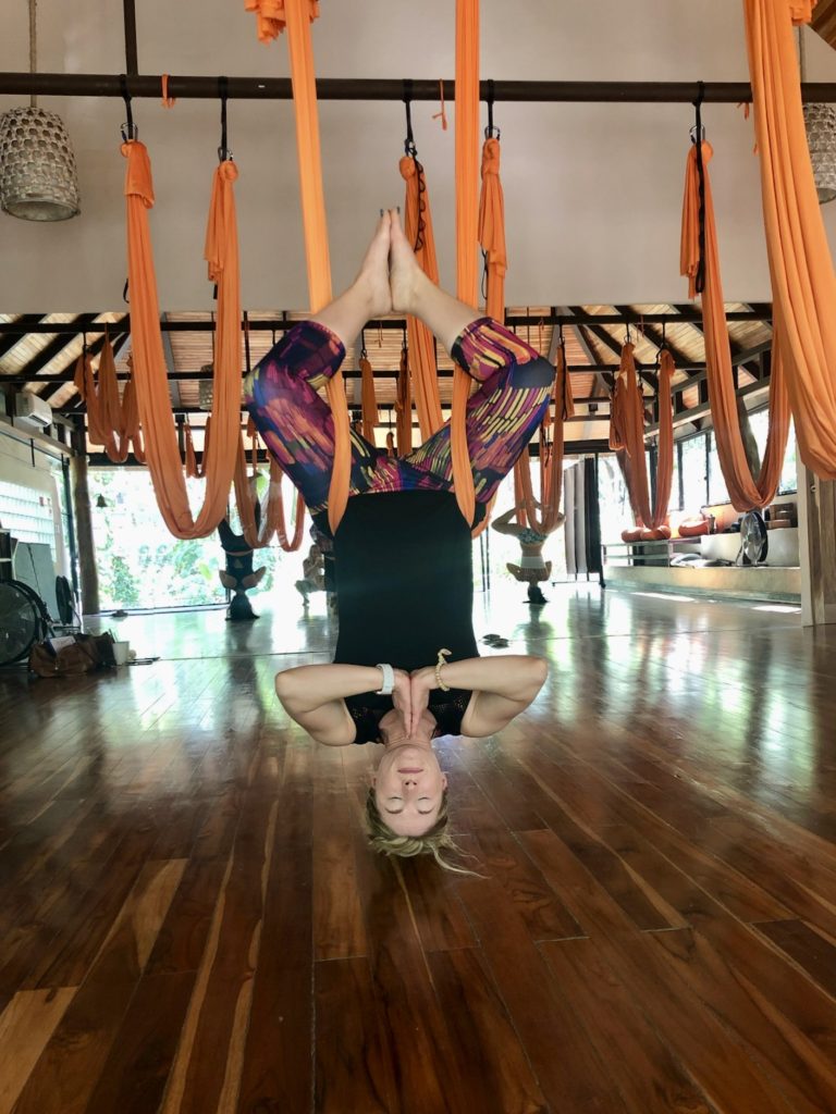 A woman practices aerial yoga by hanging upside down in a large, sunlit studio, using vibrant orange hammocks suspended from the wooden ceiling.