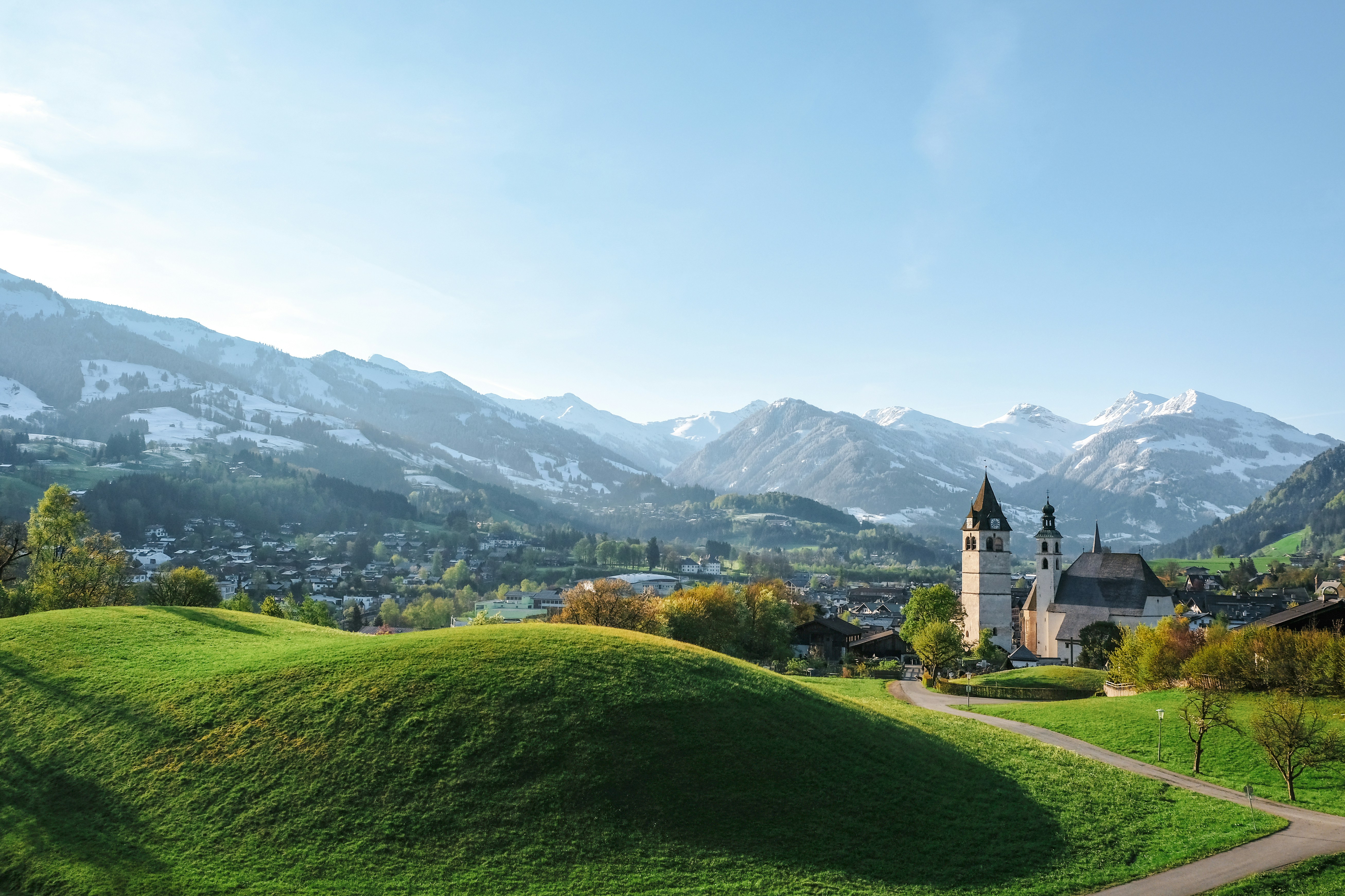 Historische Kirche auf grünem Hügel mit Blick auf schneebedeckte Alpen – harmonisches Zusammenspiel von Natur und Kultur.